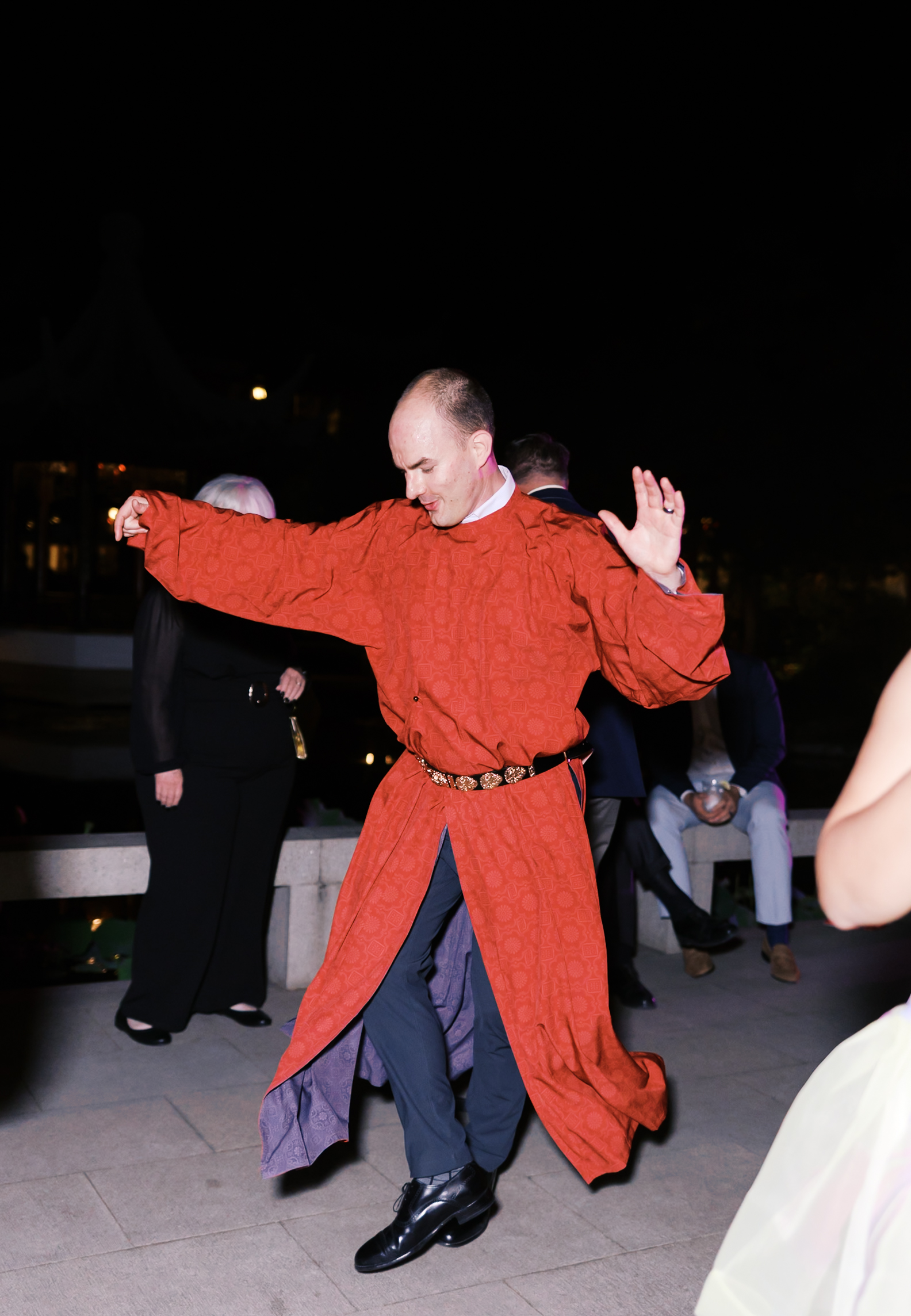 Groom dancing in traditional red Chinese wedding robe during evening reception celebration.