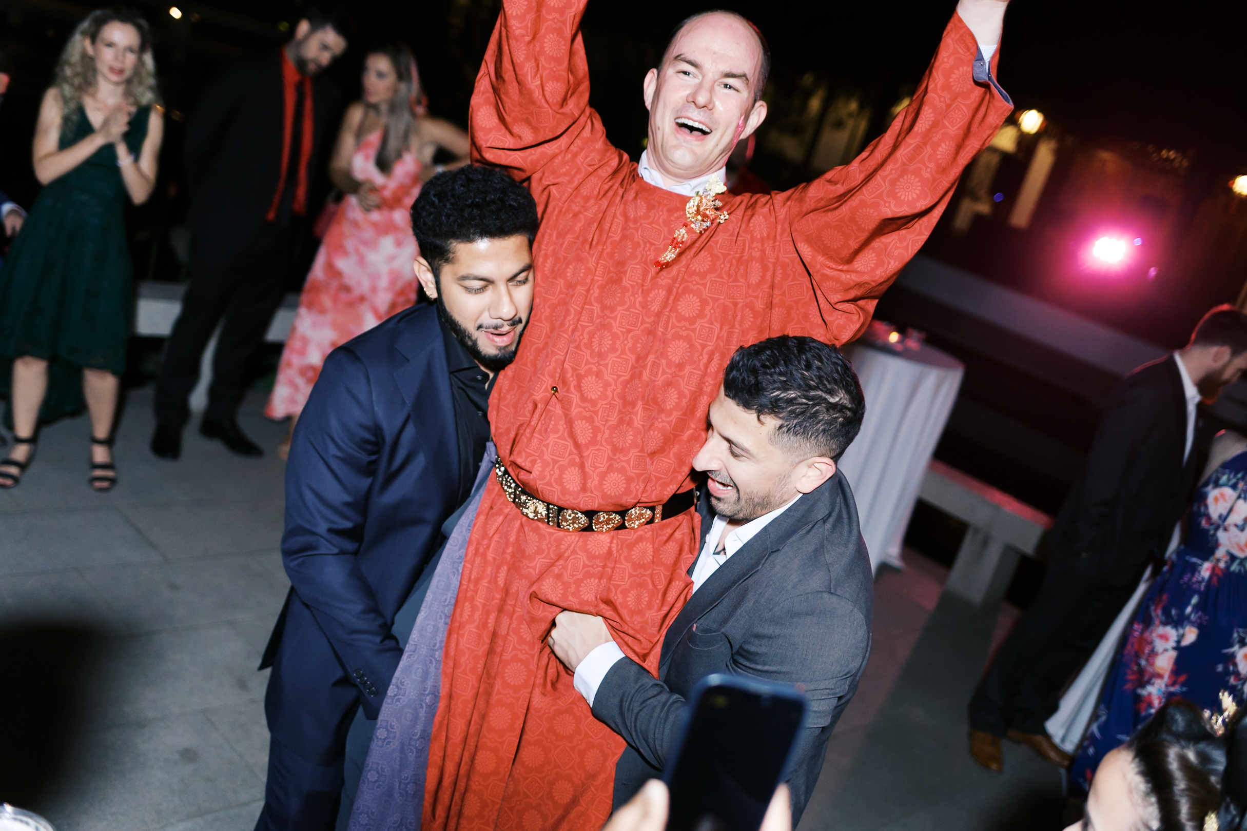 Wedding guests lift groom in traditional red Chinese attire during lively evening dance celebration.
