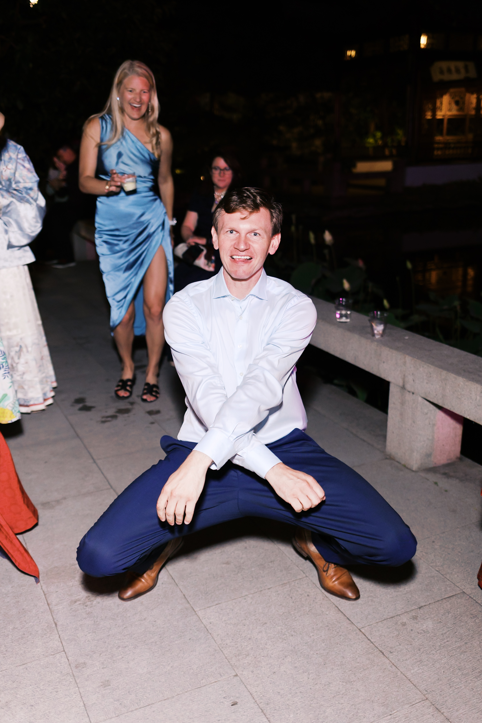 Wedding guest dancing playfully on outdoor reception floor while other guests laugh and cheer nearby.