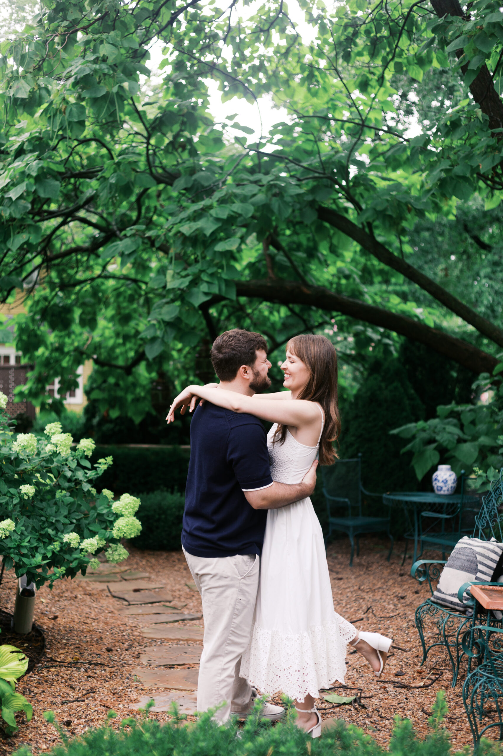 Couple embracing beneath large green trees in a quiet garden setting.