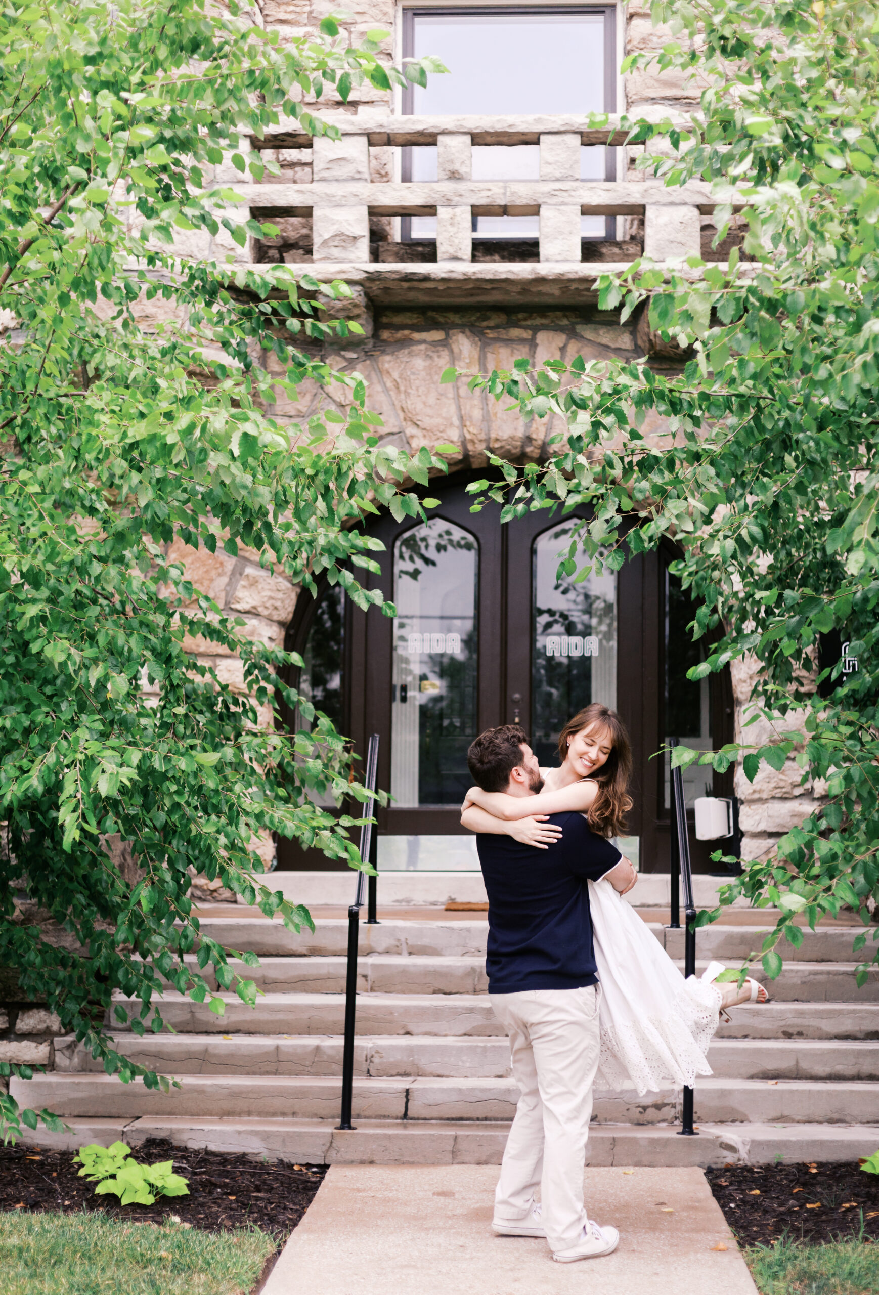 Groom lifting bride-to-be outside a stone building during a playful engagement session.