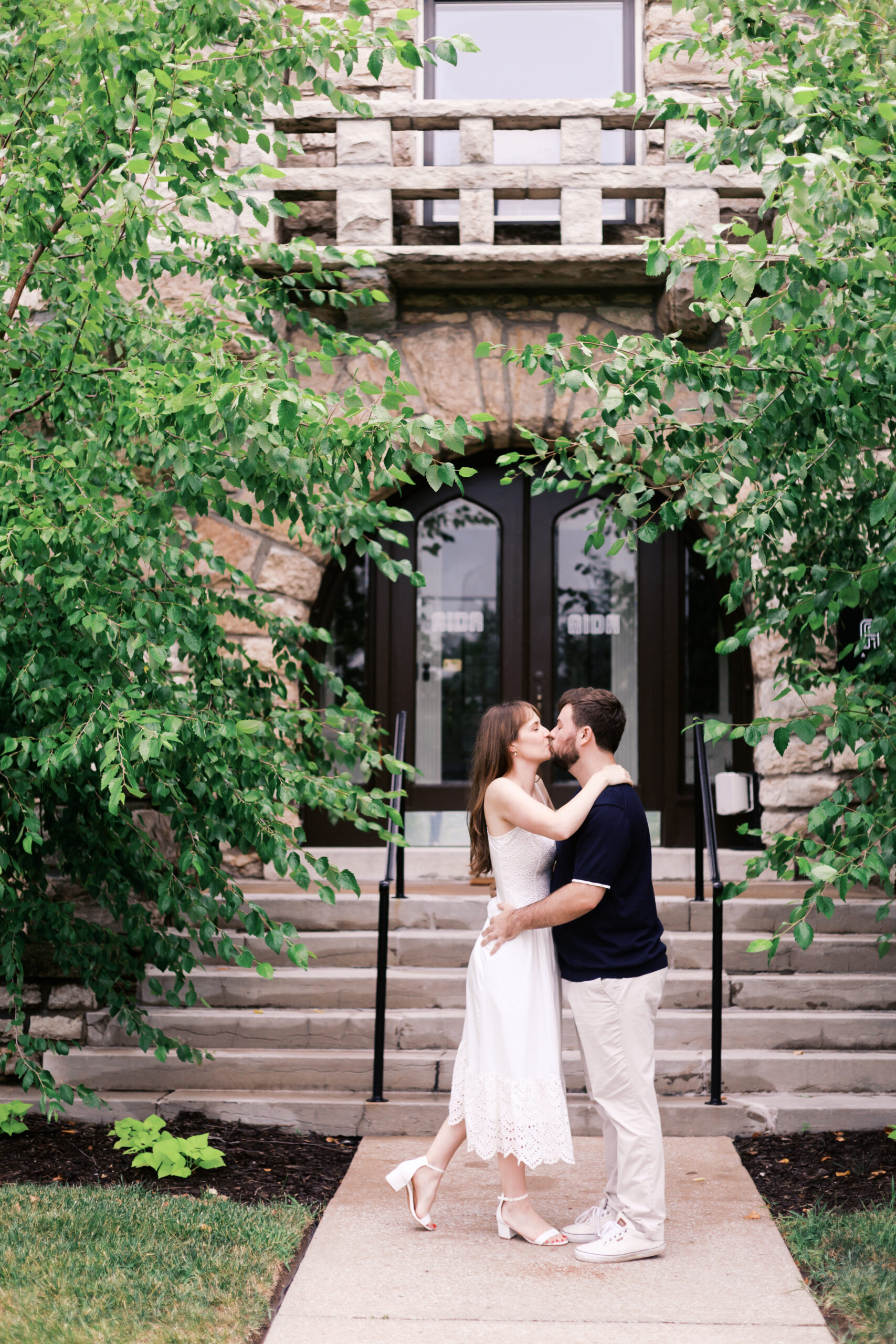 Couple kissing outside a historic entrance surrounded by leafy branches and stone architecture.
