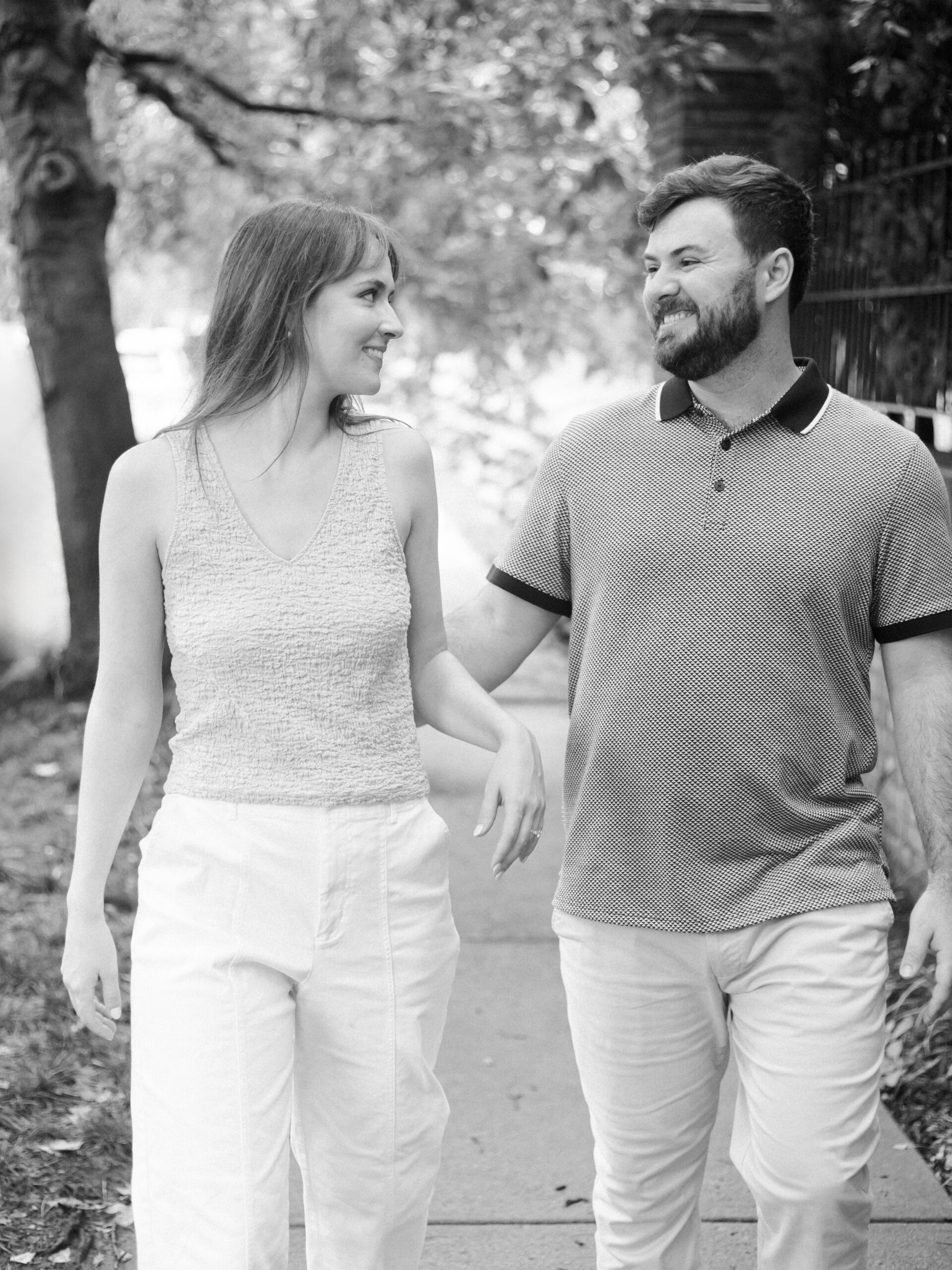Black-and-white portrait of engaged couple walking together along a tree-lined sidewalk.