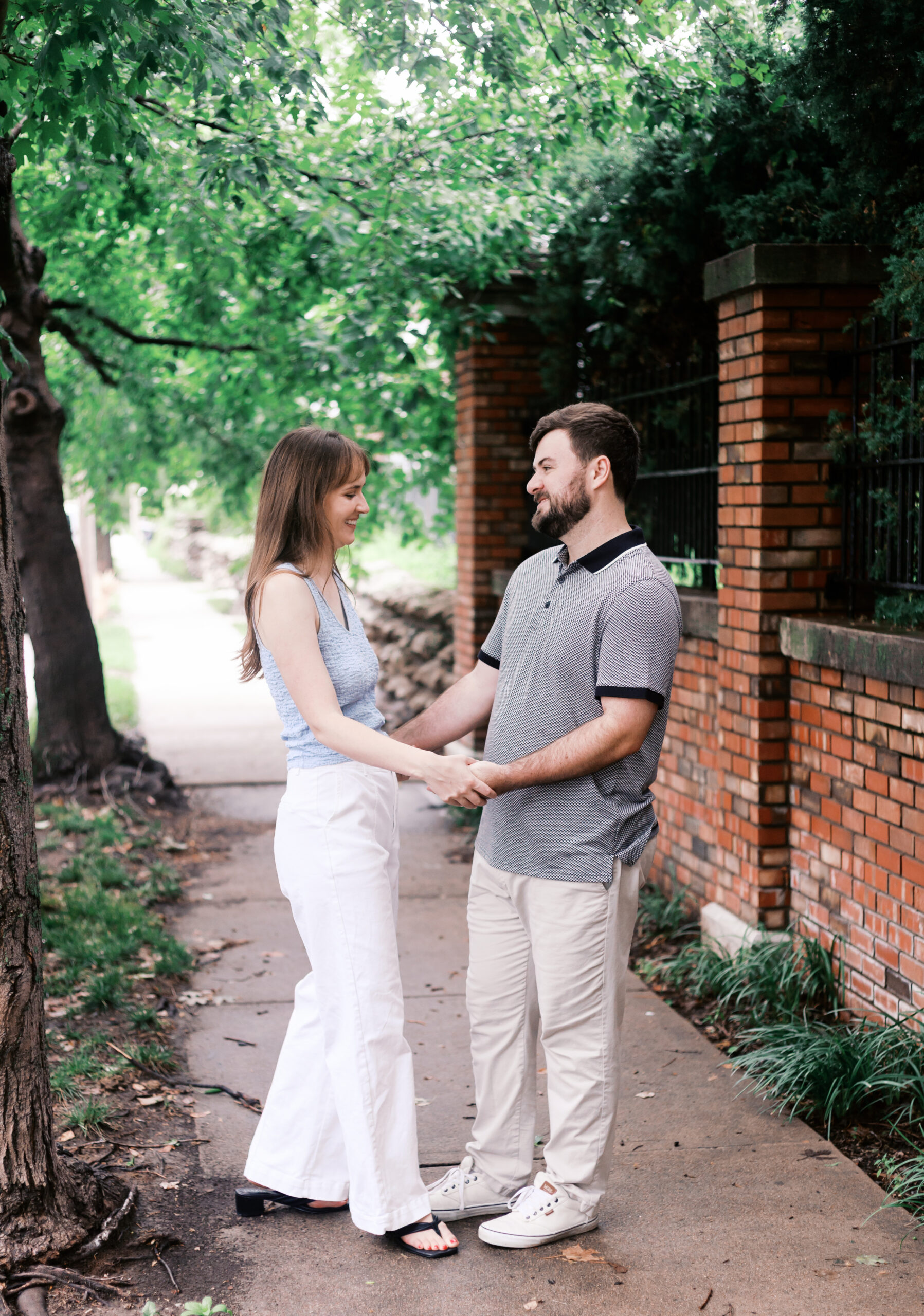 Couple holding hands beside a brick wall and shaded sidewalk during an outdoor portrait session.