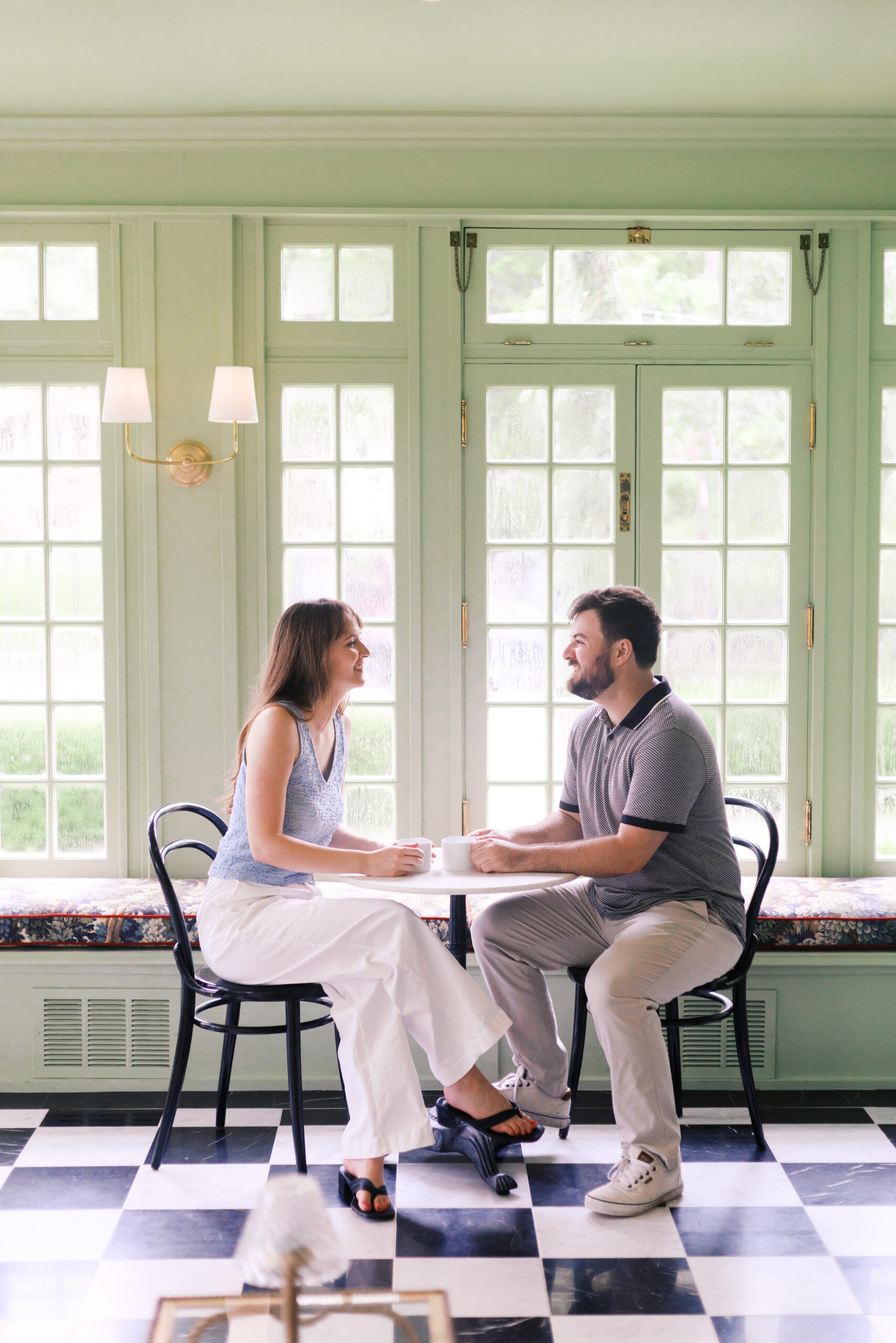 Couple sharing coffee at a small café table inside a bright sunroom.