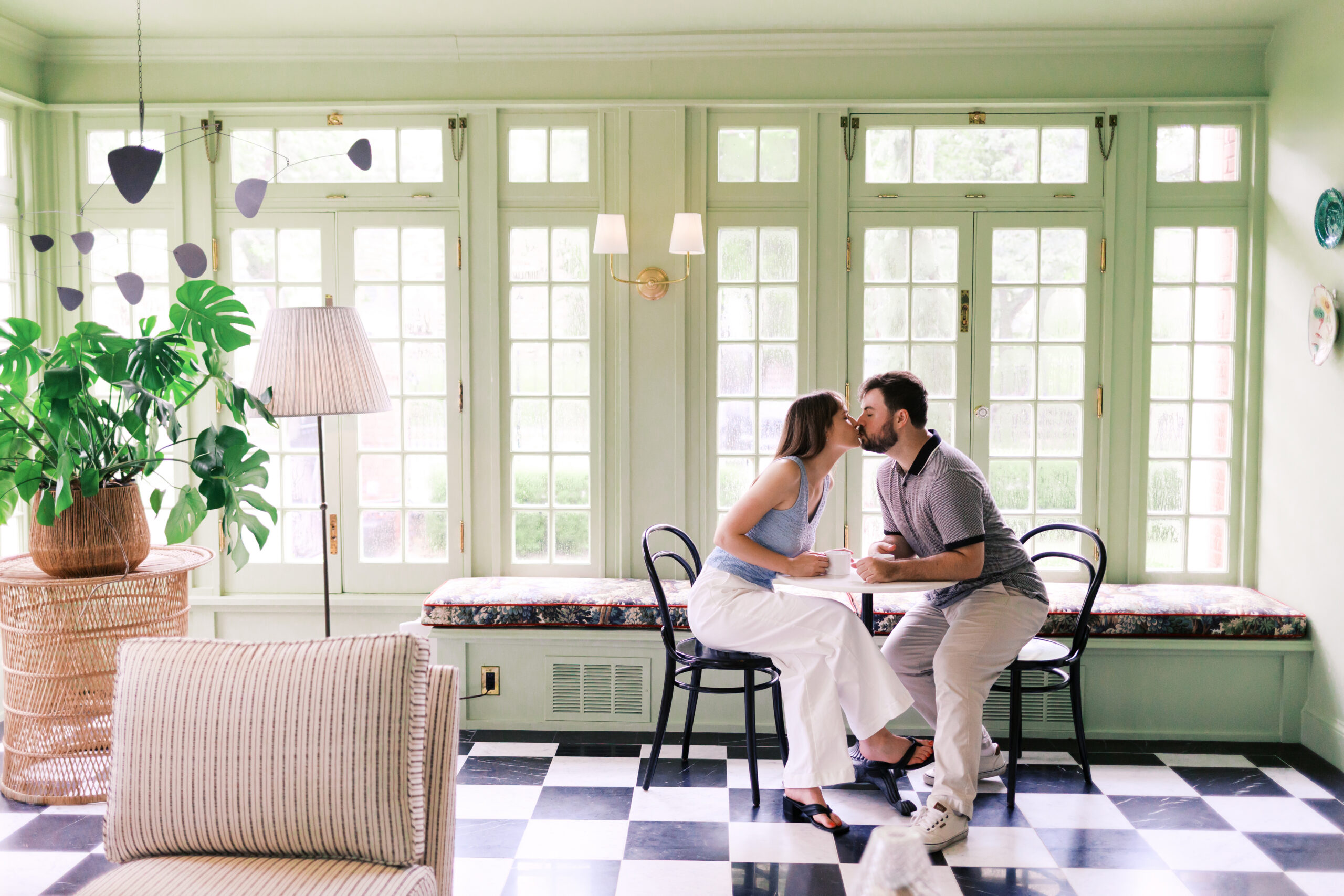 Couple kissing across a café table in a bright room with mint green walls and checkered floors.