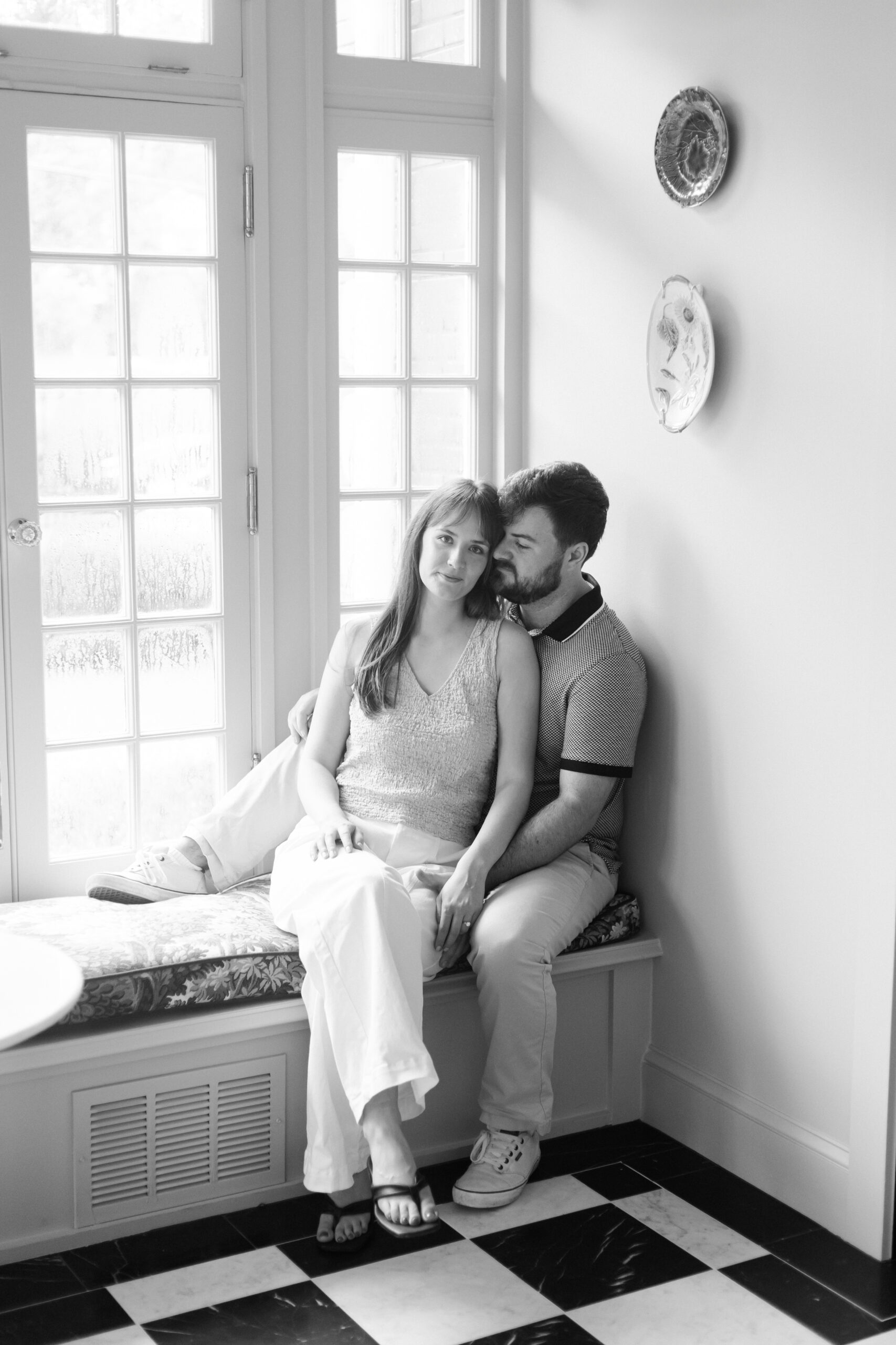 Black-and-white portrait of engaged couple cuddling by a window seat indoors.