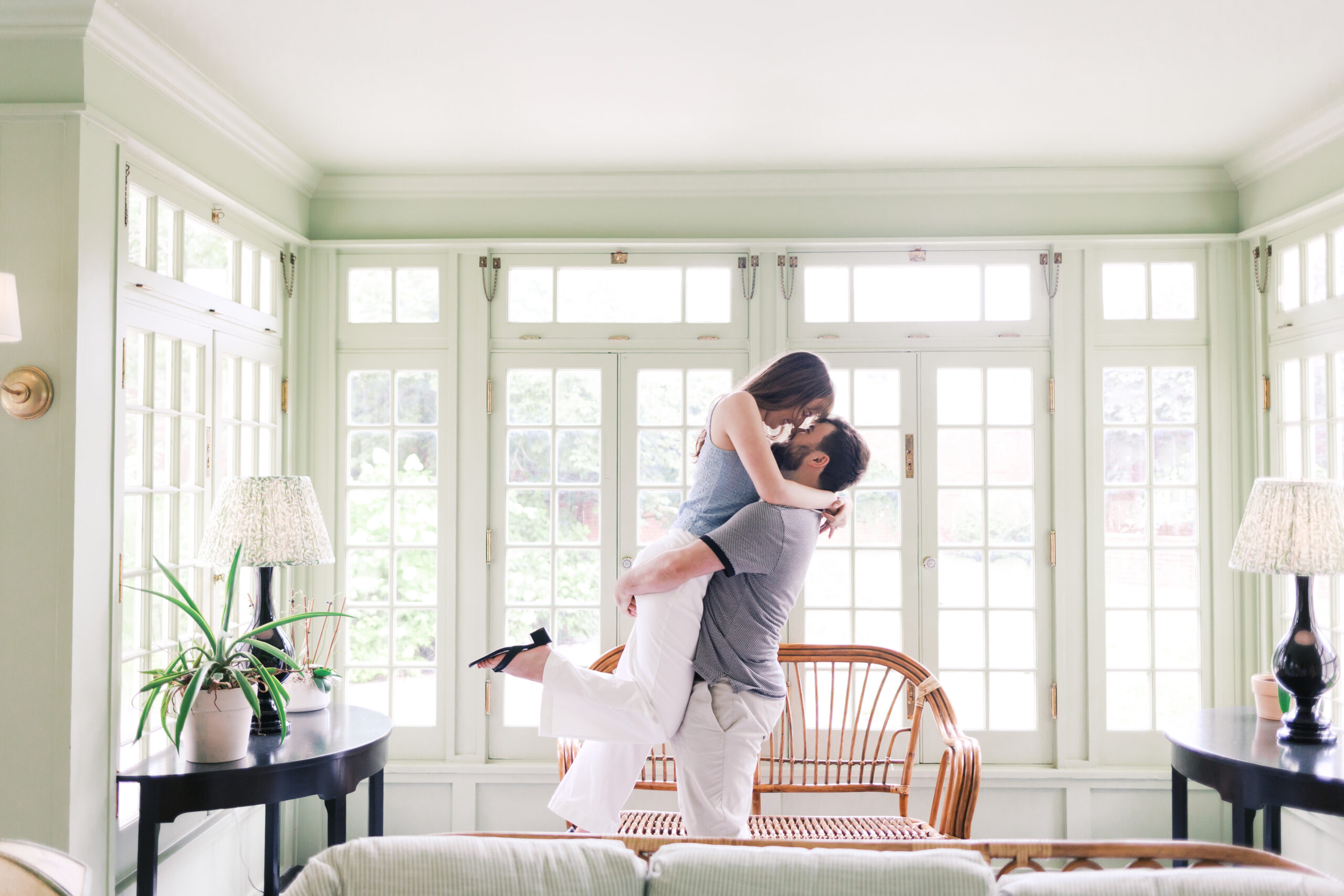Groom lifting bride-to-be in a bright sunroom filled with natural light and vintage details.