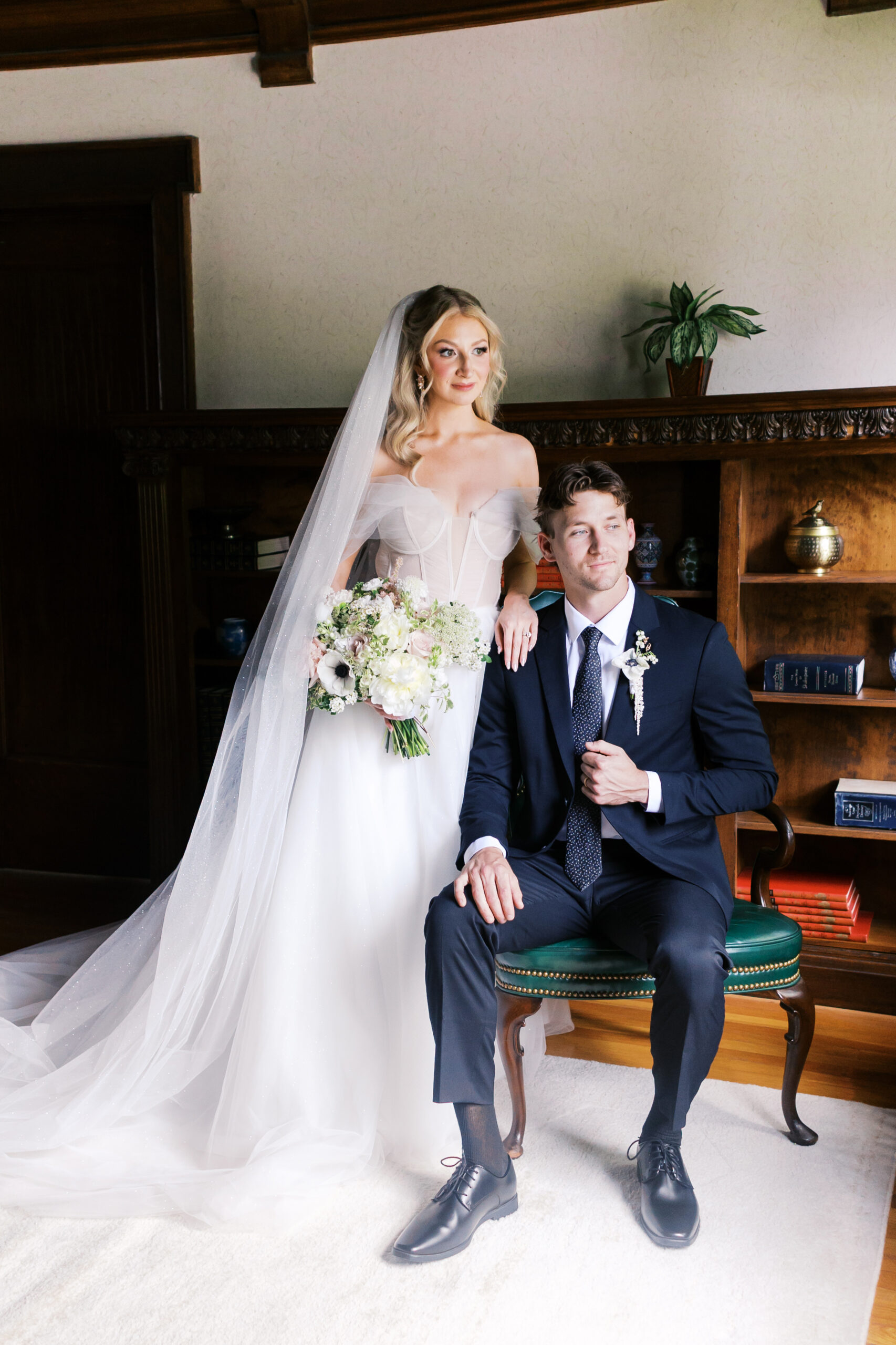 Bride in an off-the-shoulder gown standing beside groom in a navy suit inside Simpson House.