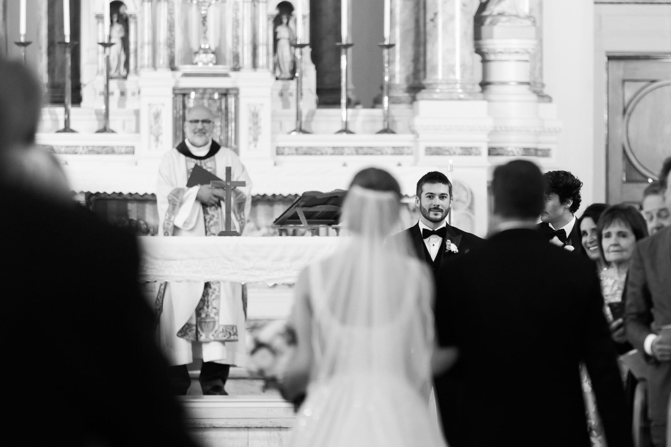 Groom watching the bride walk down the aisle during a black and white church wedding ceremony.