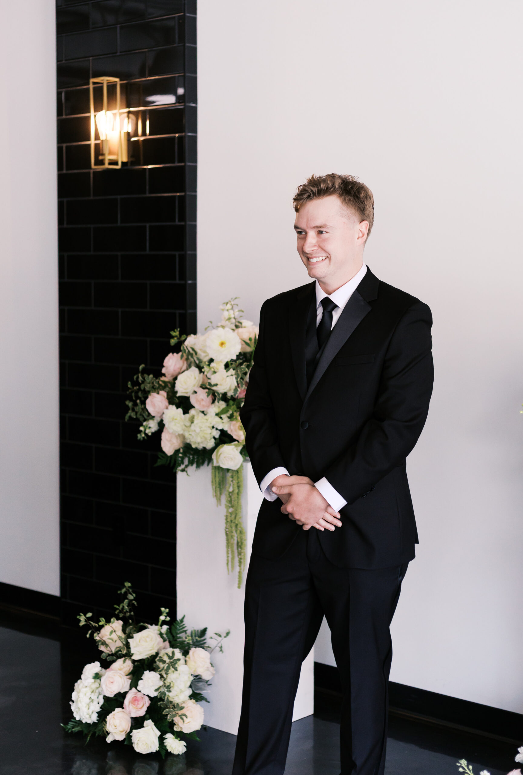 Groom in a black suit standing near floral arrangements before an indoor wedding first look.