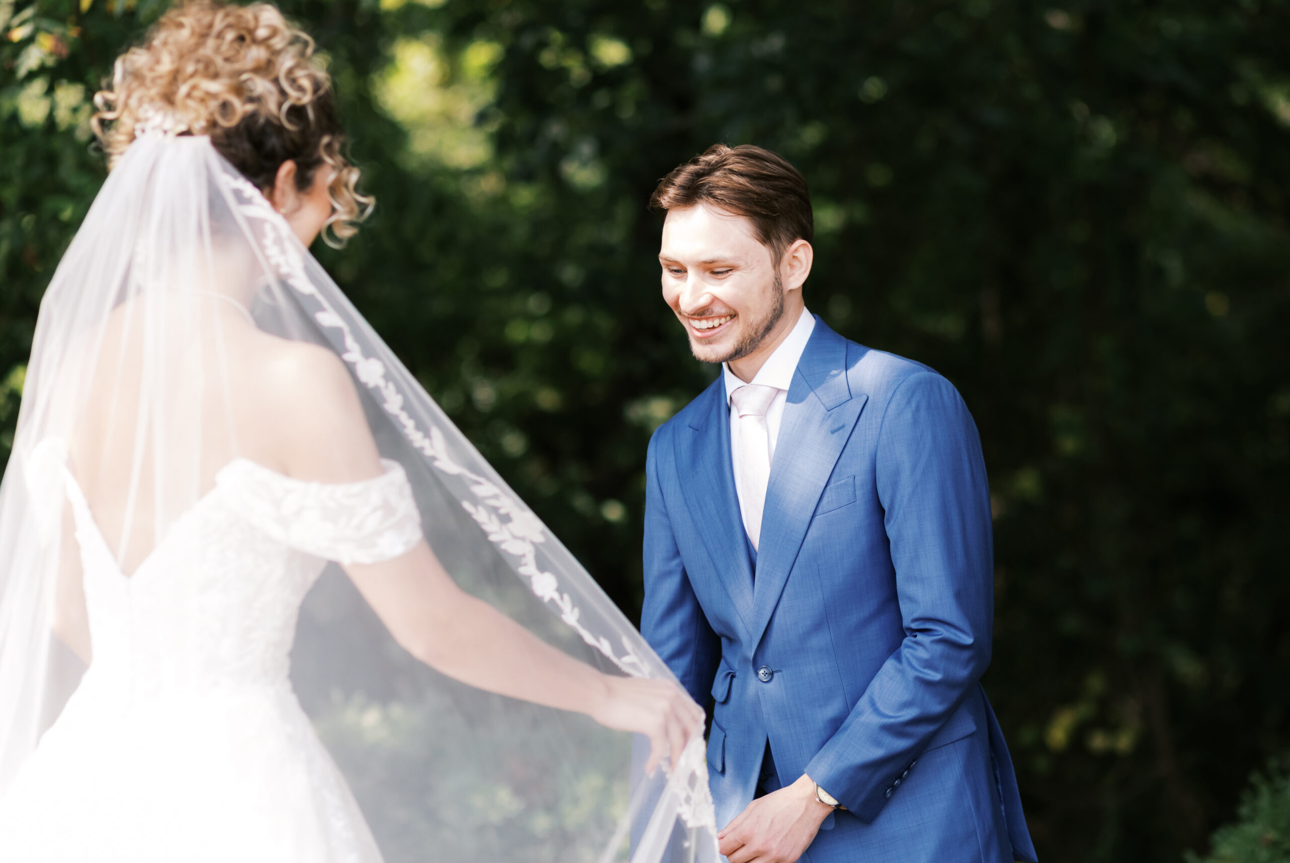 Groom in a blue suit smiling during an outdoor first look with the bride.