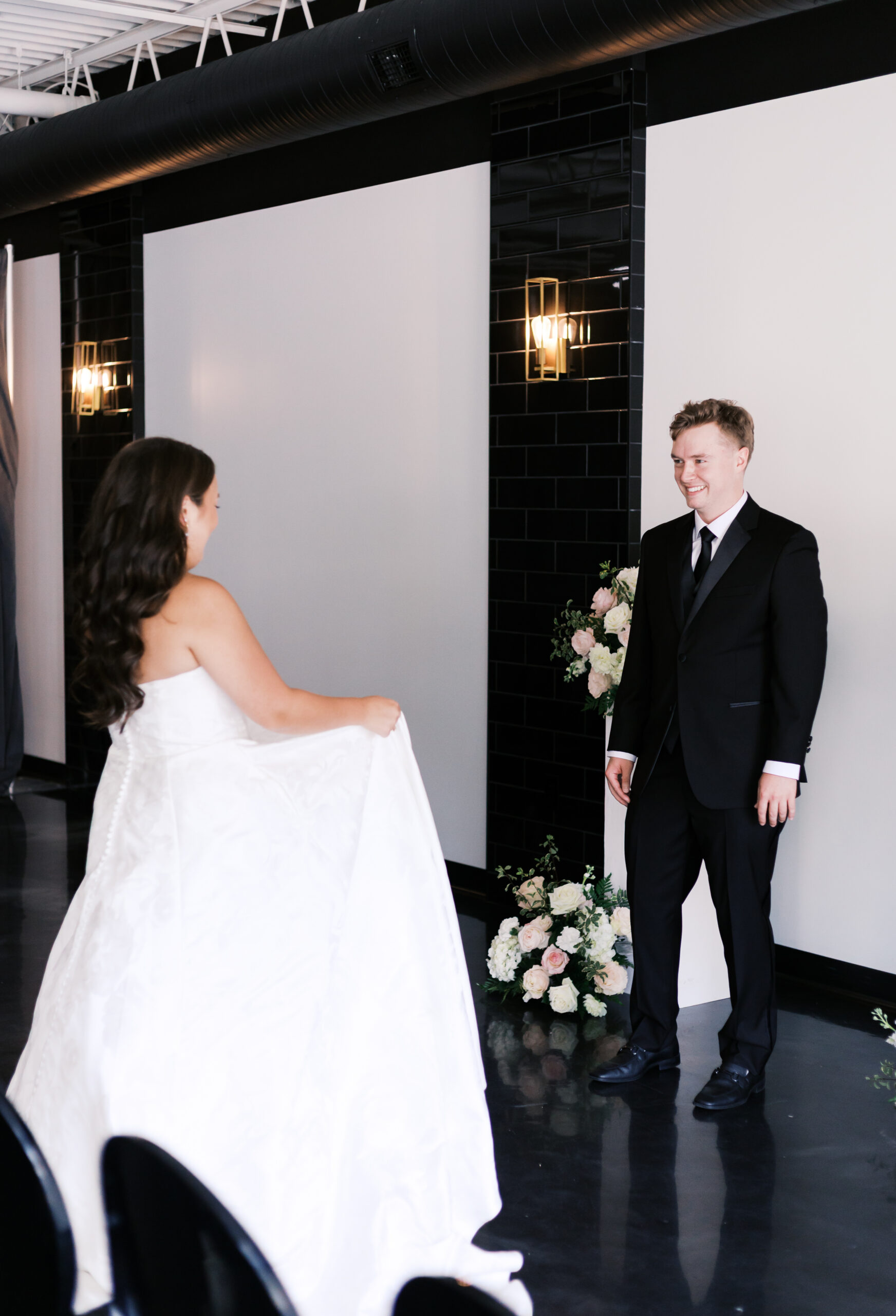 Bride walking toward the groom during an indoor first look with modern black tile and floral decor.