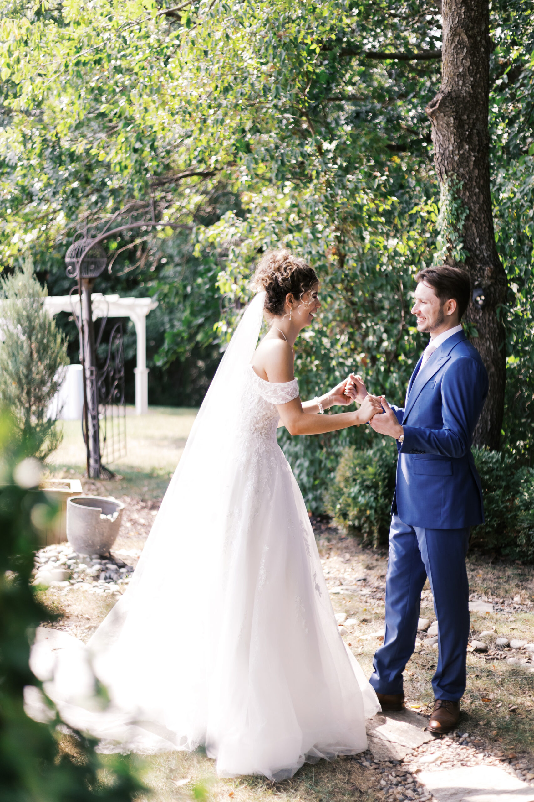 Bride and groom holding hands during an outdoor first look beneath leafy trees.