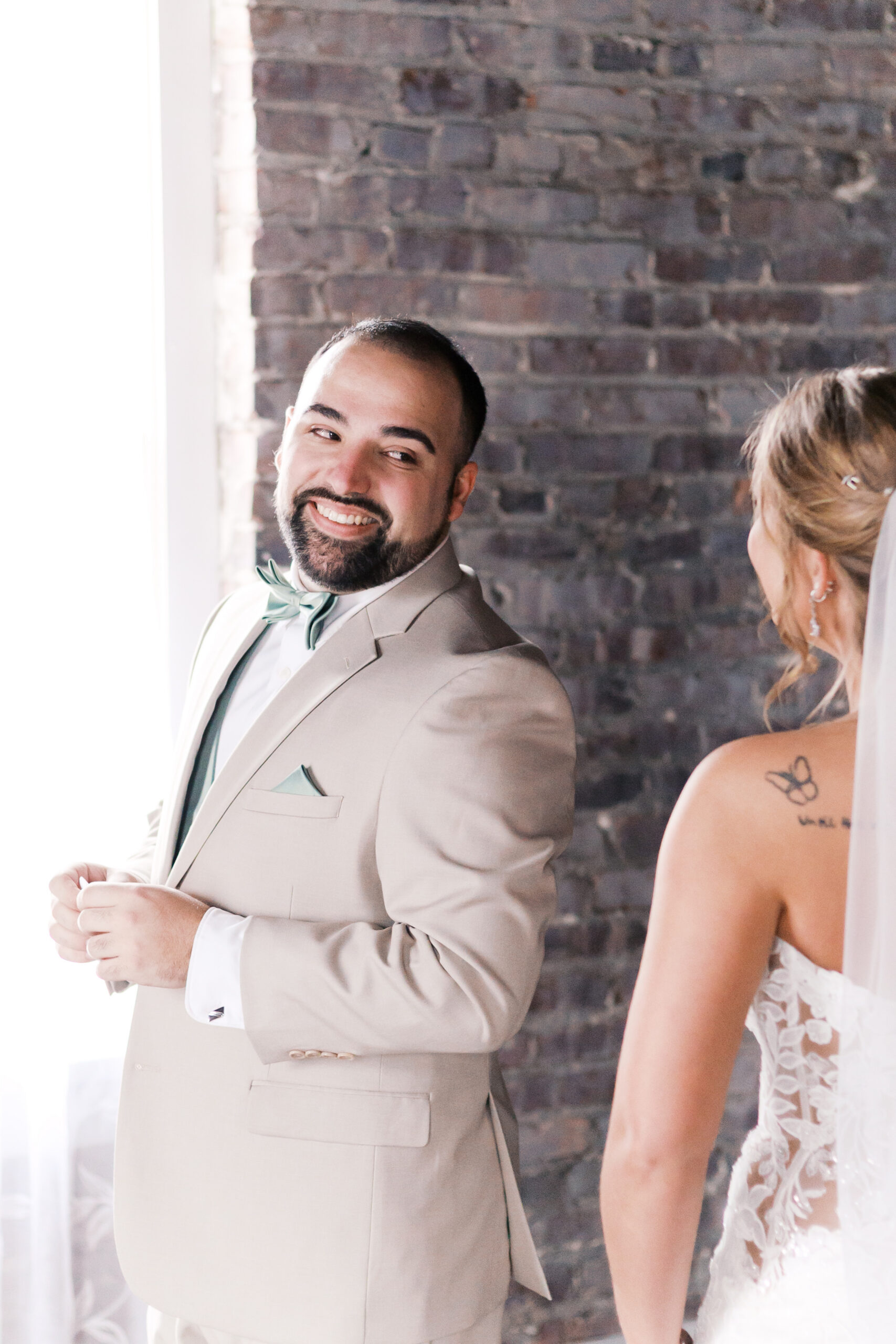 Groom in a beige suit smiling at the bride during an indoor wedding first look.
