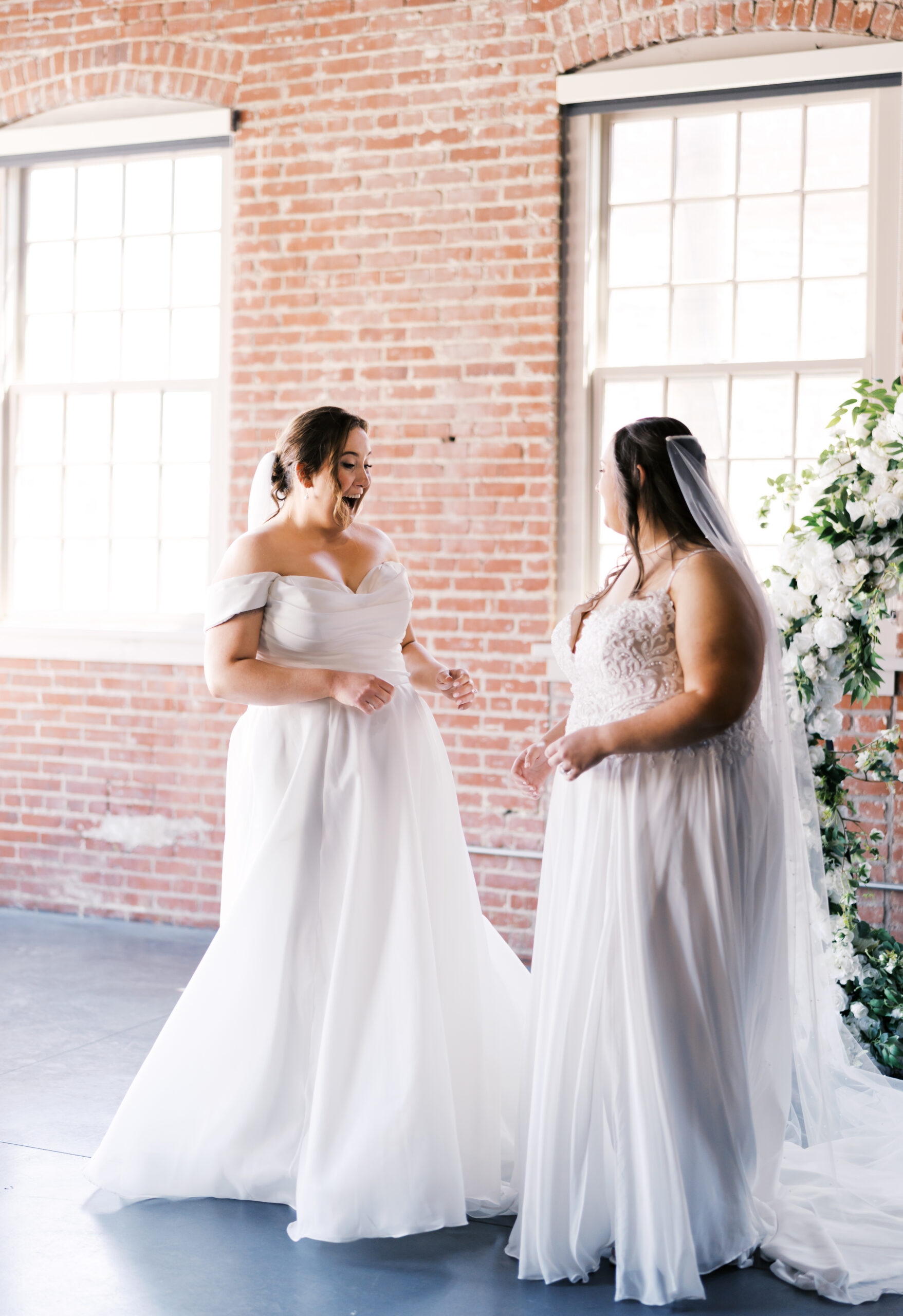 Two brides laughing together in white wedding dresses inside a bright brick-walled venue.
