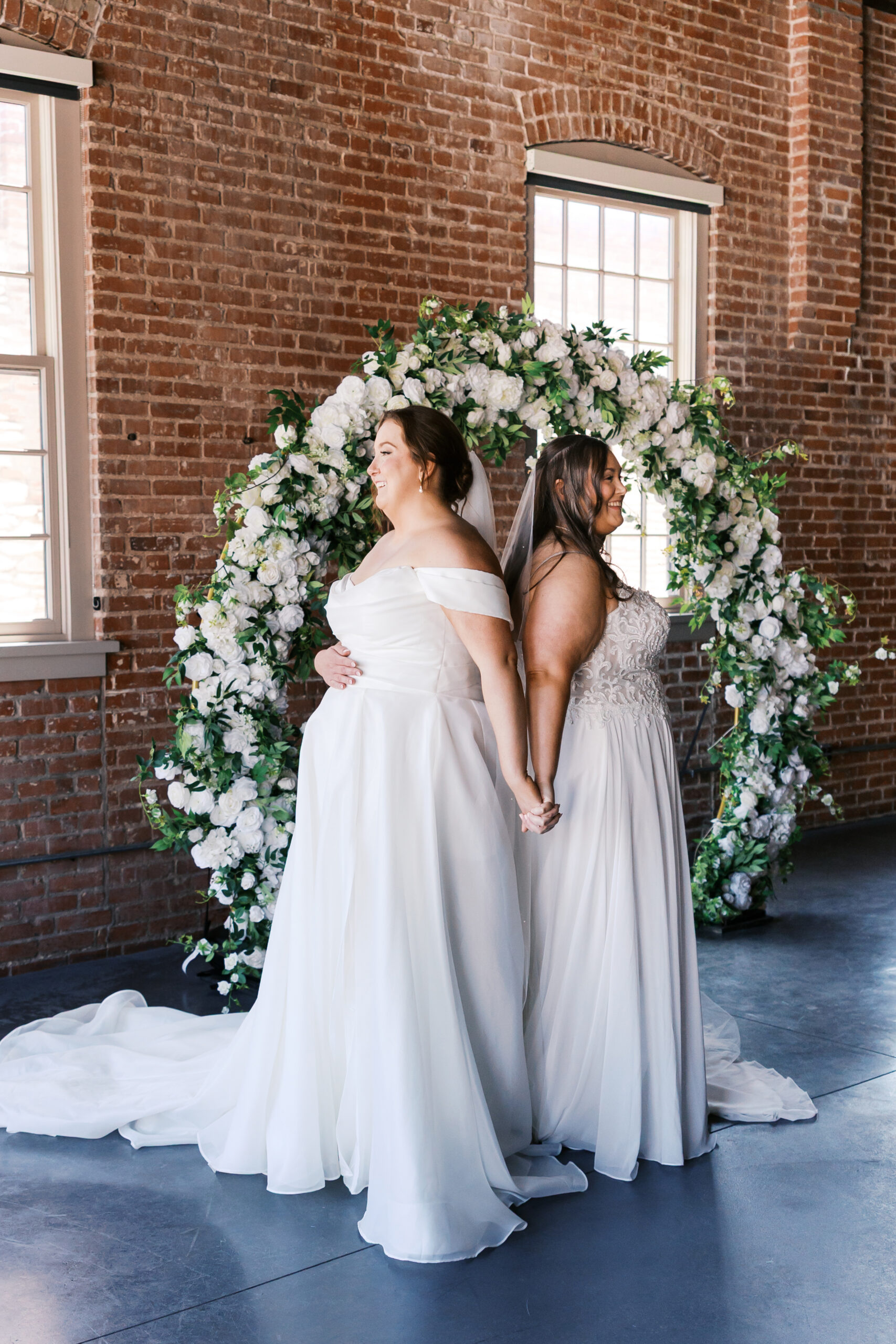 Two brides standing back to back and holding hands in front of a white floral arch and brick wall.