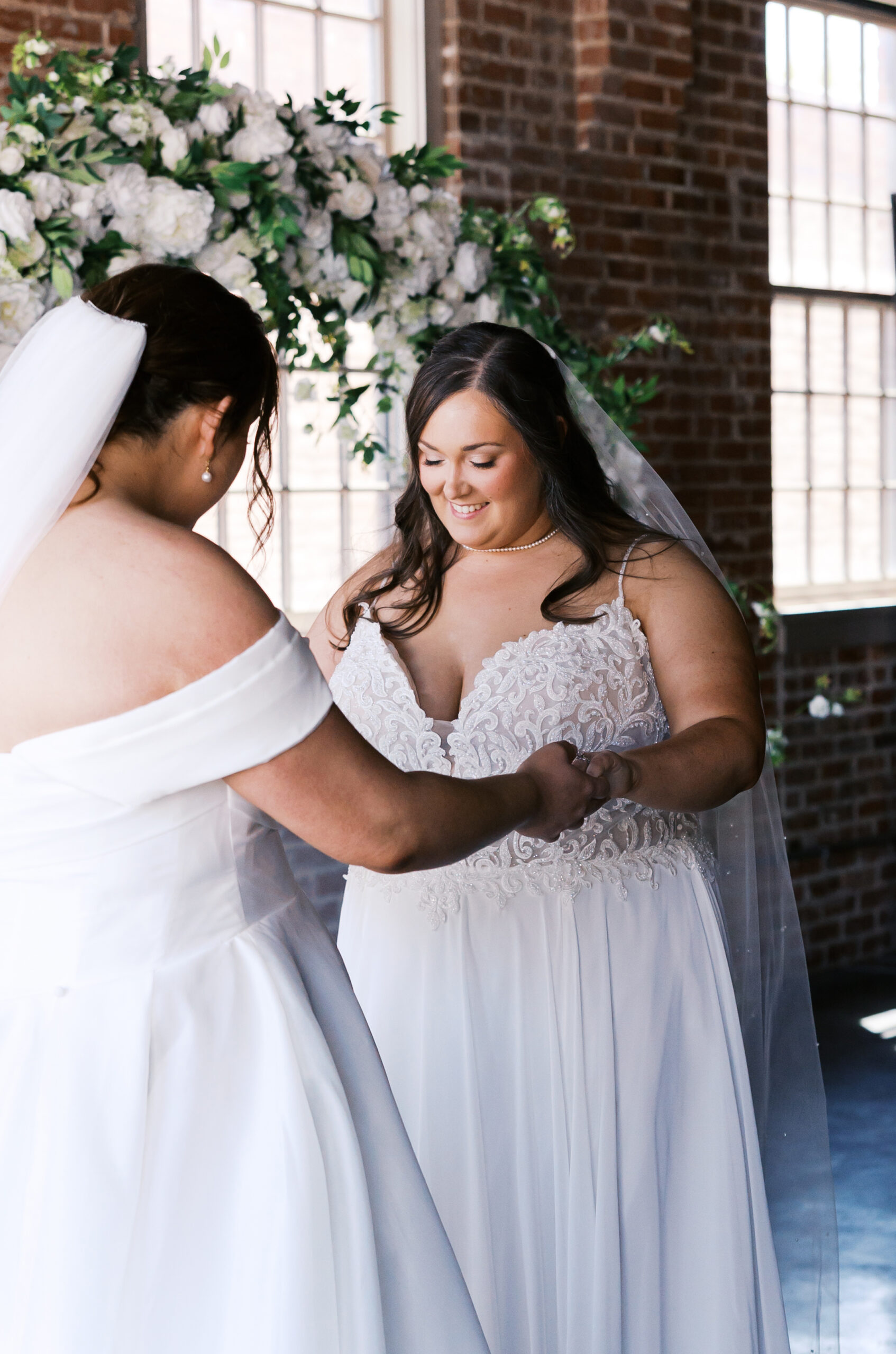 Two brides holding hands during a first look inside a brick wedding venue.