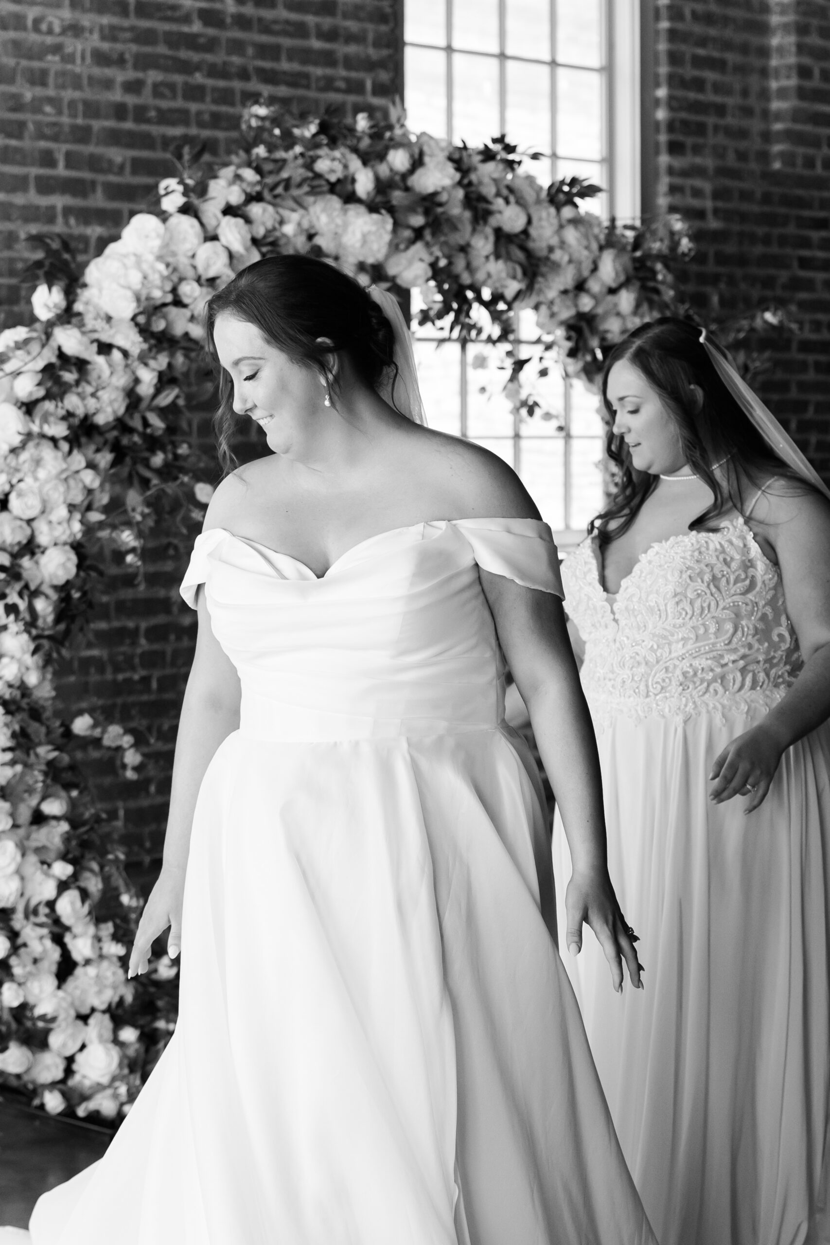 Two brides in white wedding dresses standing near a floral arch in a black and white portrait.