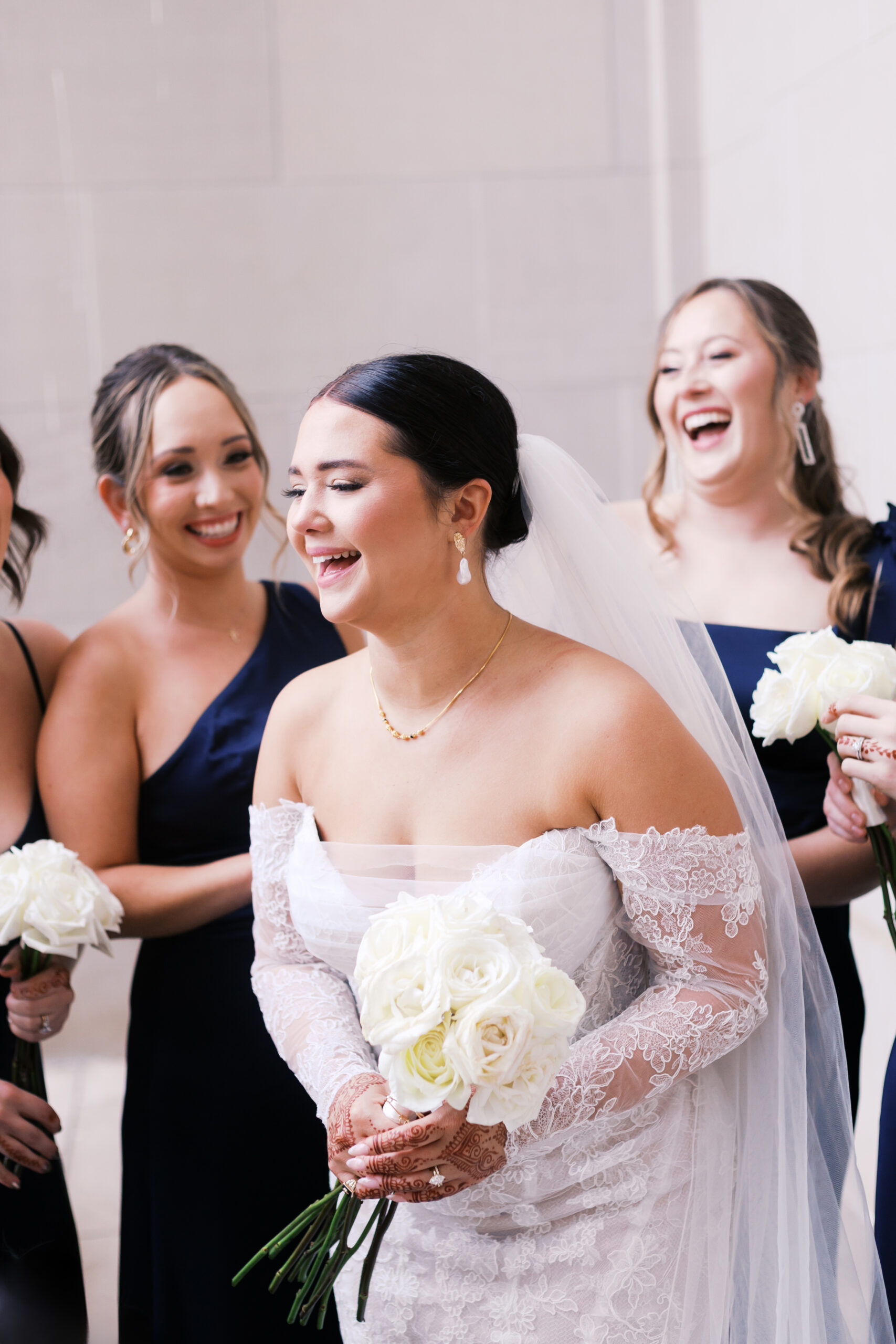 Bride laughs with bridesmaids while holding white roses on her wedding day.