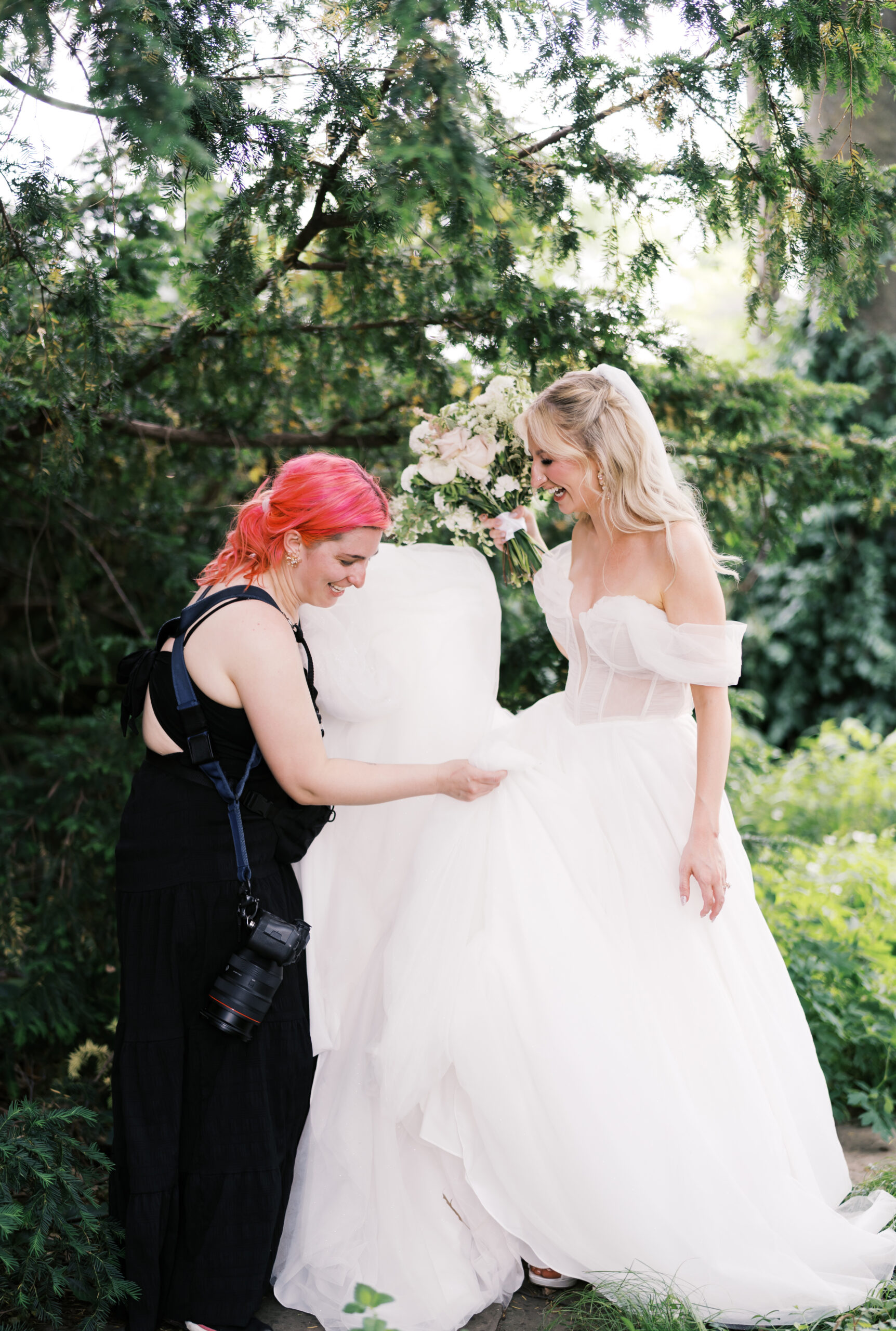 Photographer fluffs a bride’s tulle wedding dress during portraits in a leafy garden.