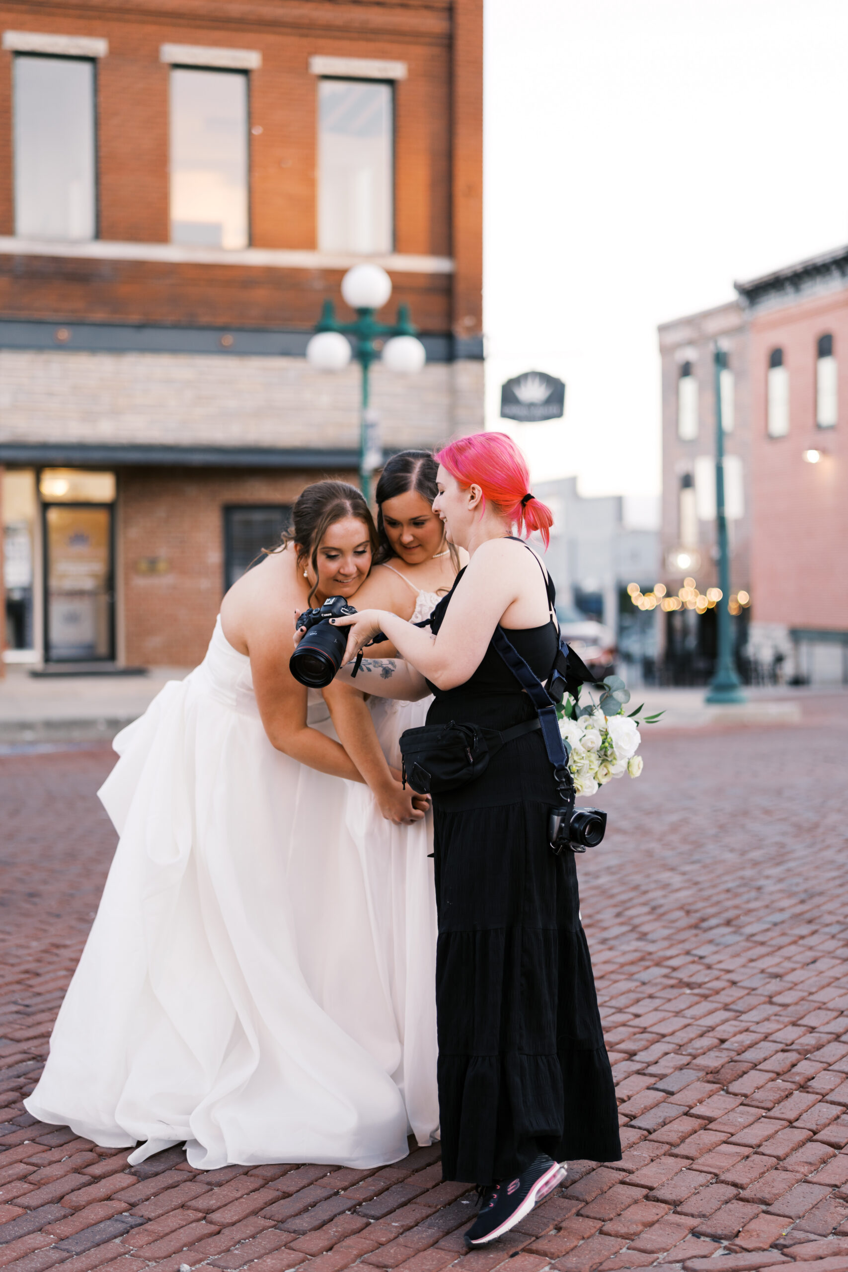 Photographer shows two brides a camera preview on a brick street.