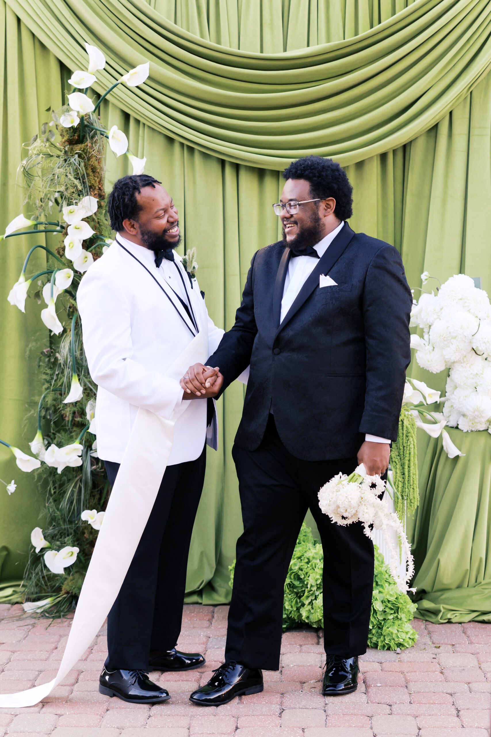 Two grooms hold hands before a green draped ceremony backdrop with white florals.