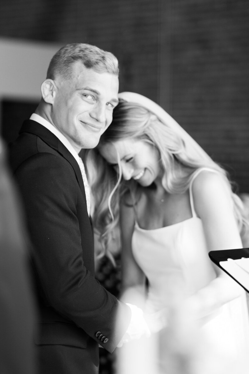 Groom smiling beside bride in candid black-and-white wedding portrait during indoor ceremony preparations.
