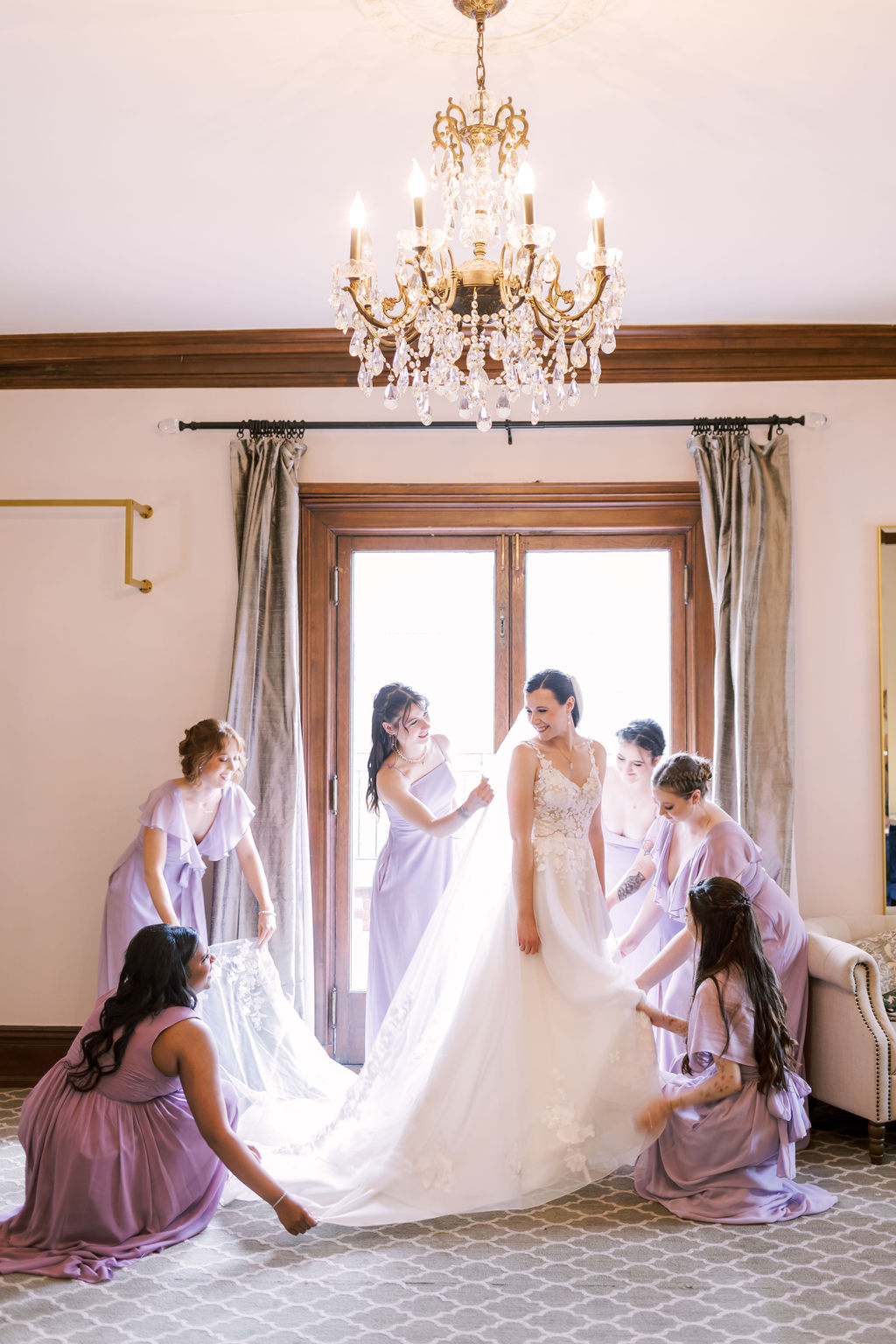 Bridesmaids help adjust the bride's wedding dress and veil in a bright bridal suite at Loose Mansion