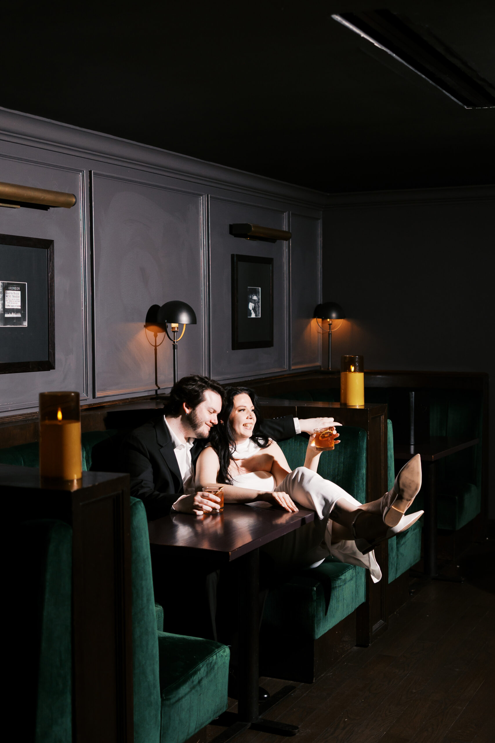 Couple relaxing together in a velvet booth, laughing and holding cocktails in a moody lounge.