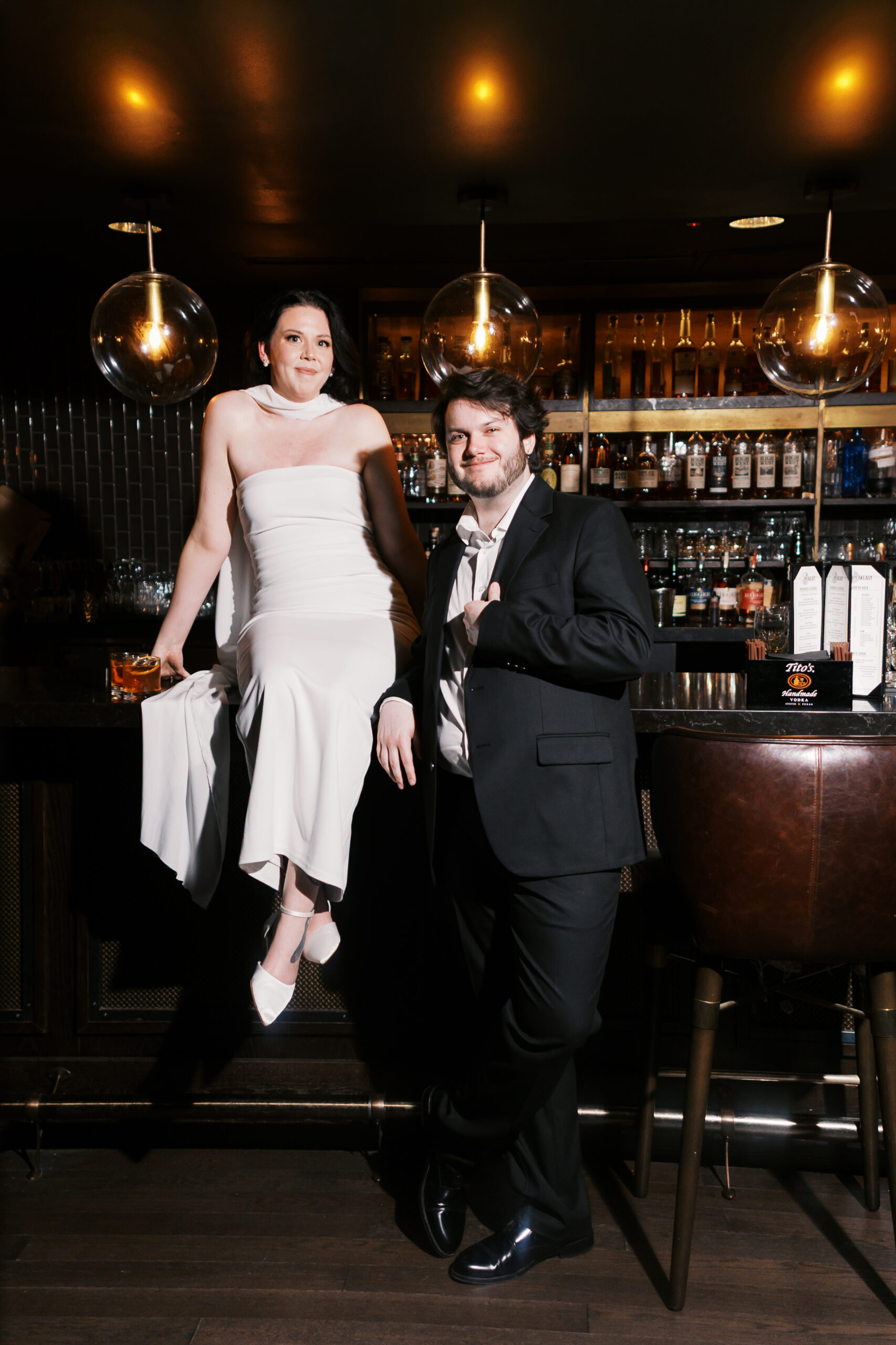 Couple dressed in formal attire posing together at the hotel bar with drinks.
