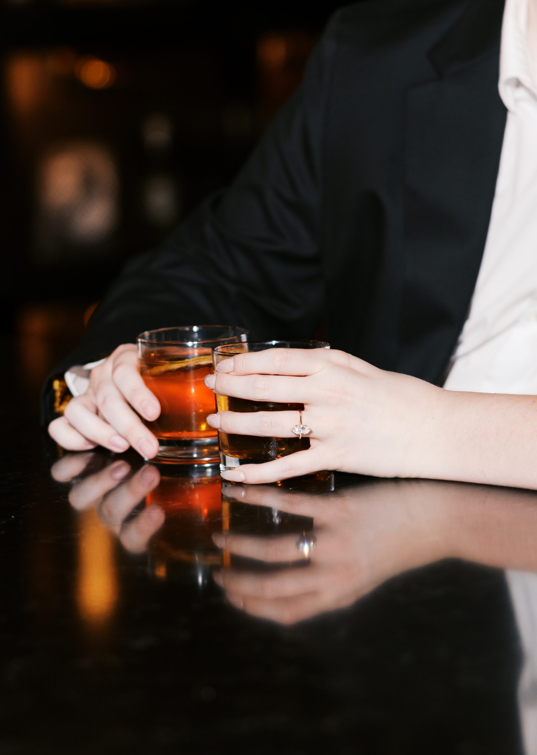 Close-up of hands holding cocktails, highlighting the engagement ring and drink reflections.