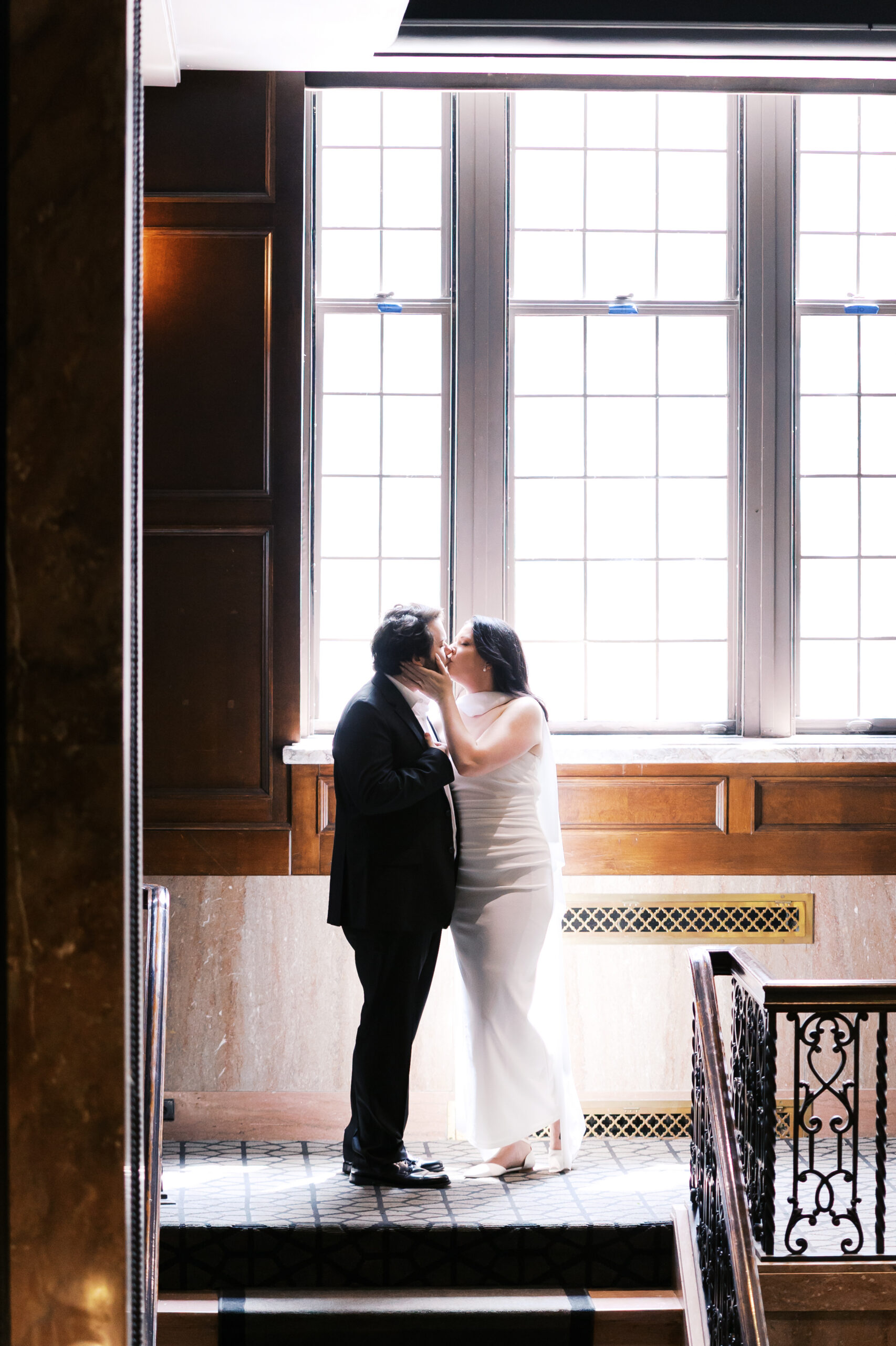 Couple sharing a kiss in front of tall windows with soft natural light.