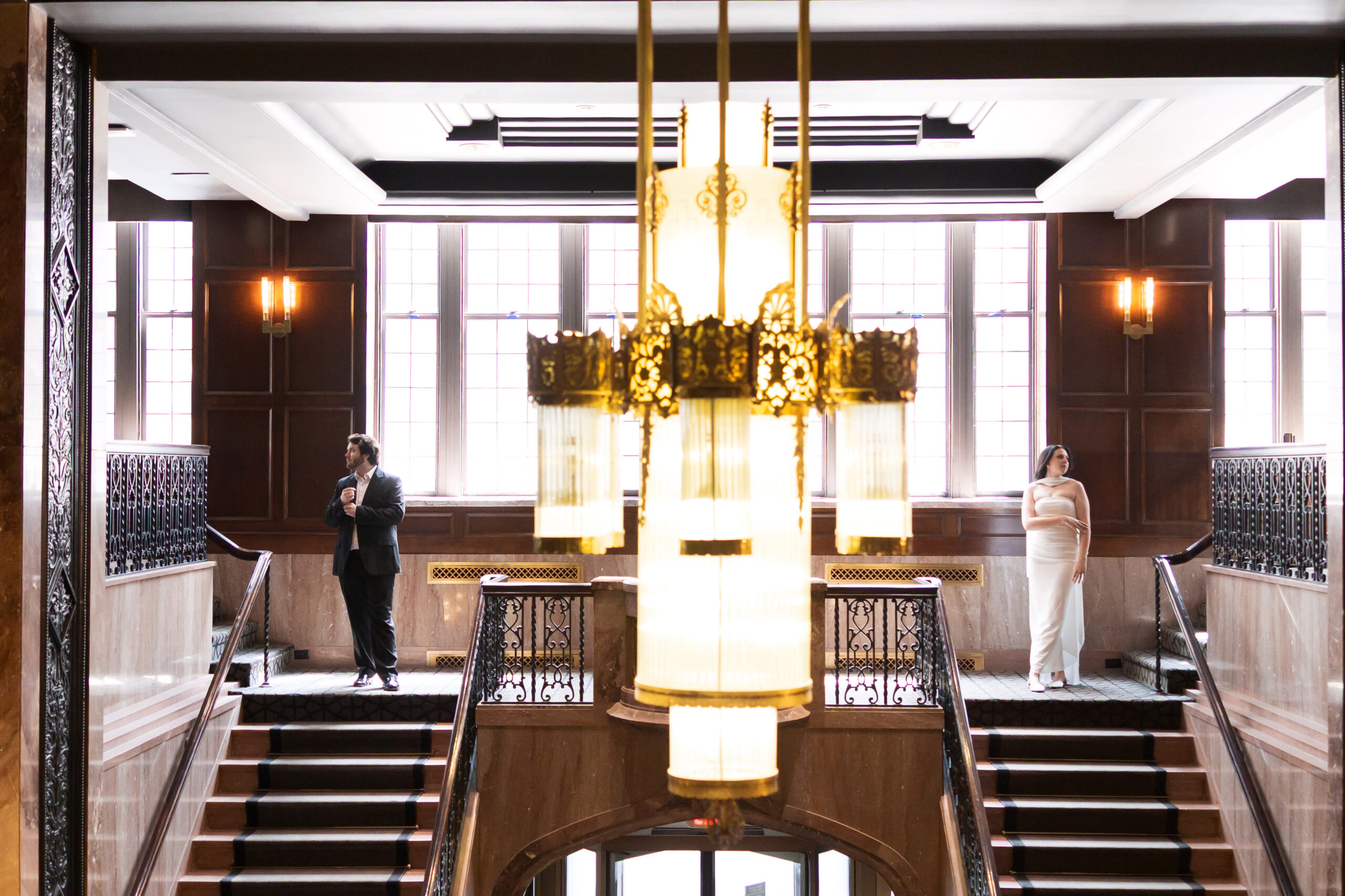 Couple standing apart across the staircase landing, framed by a vintage chandelier.