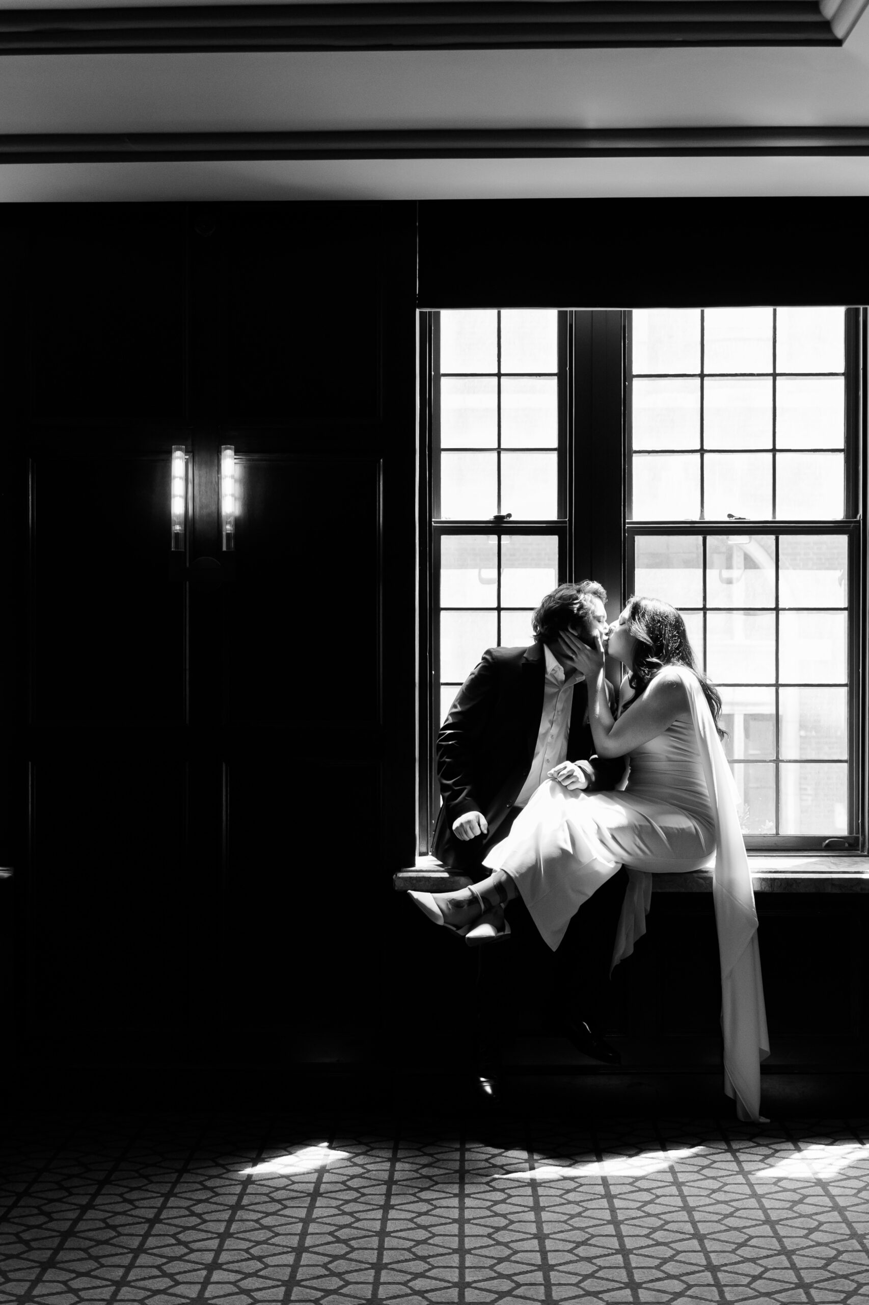 Black-and-white portrait of the couple kissing while seated on a window ledge, backlit by natural light.
