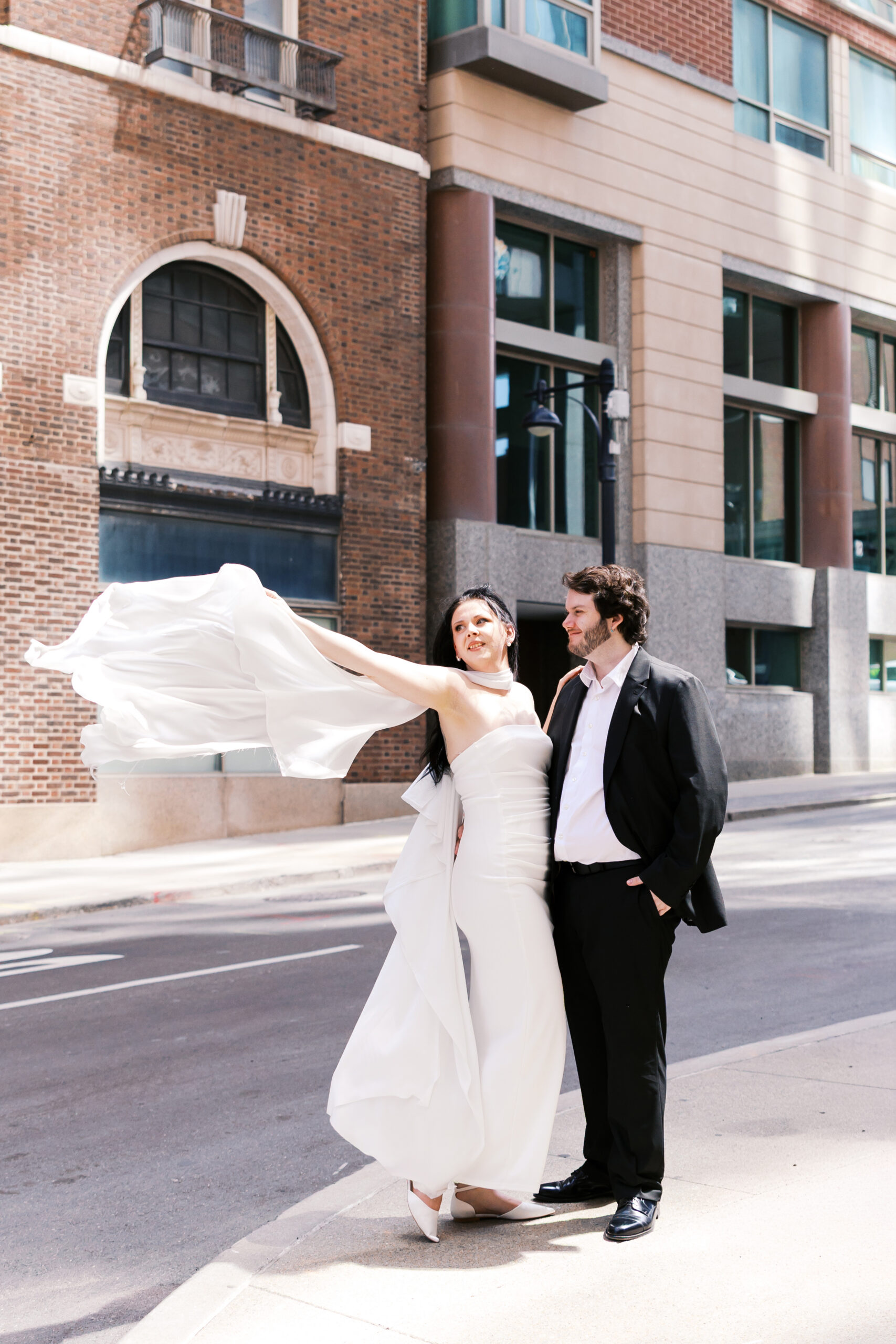 Couple posing on a downtown Kansas City sidewalk as the white scarf of the dress catches the breeze.