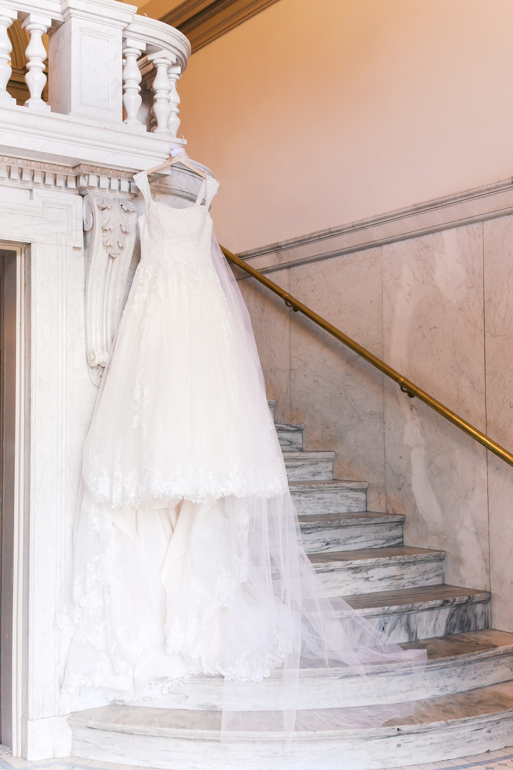 Lace wedding dress with a long veil hanging from a marble staircase inside The Loretto before the ceremony.