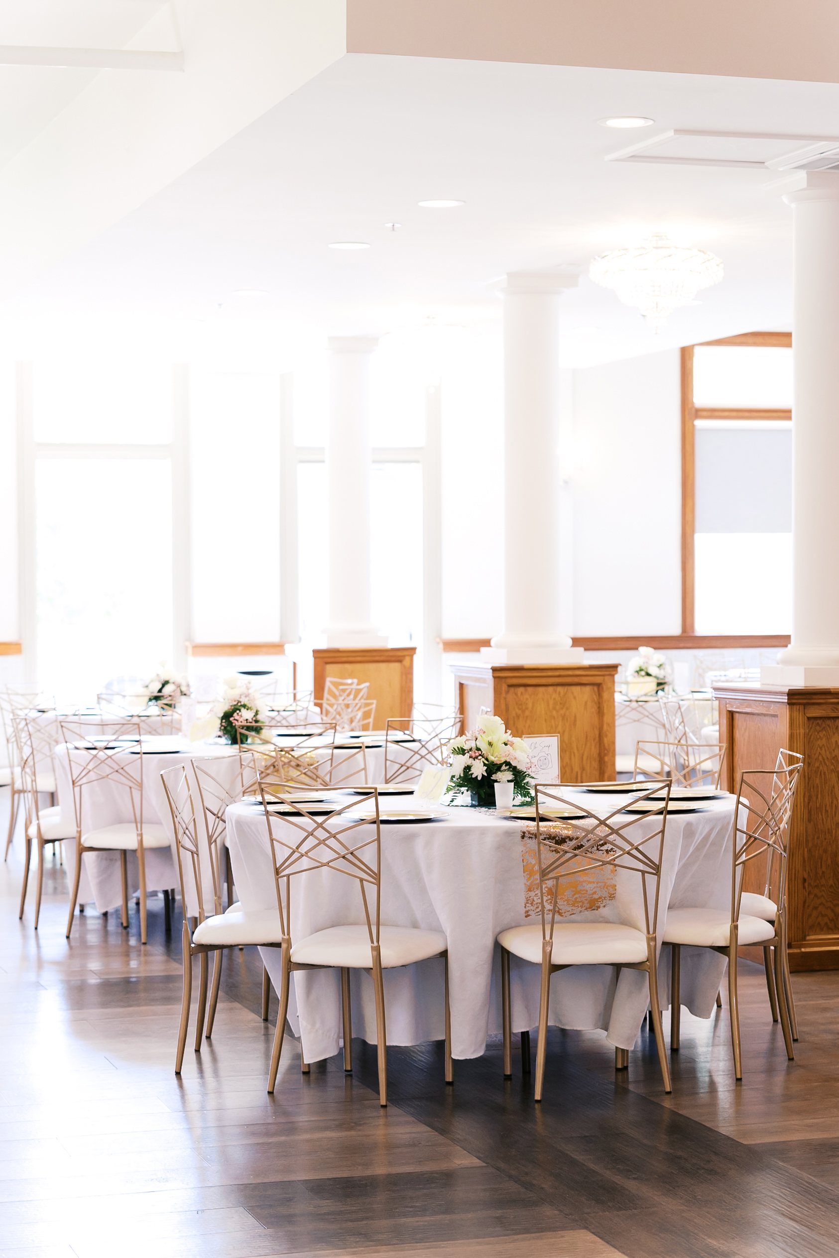 Wedding reception tables with white linens, gold chairs, and floral centerpieces inside The Loretto reception hall.