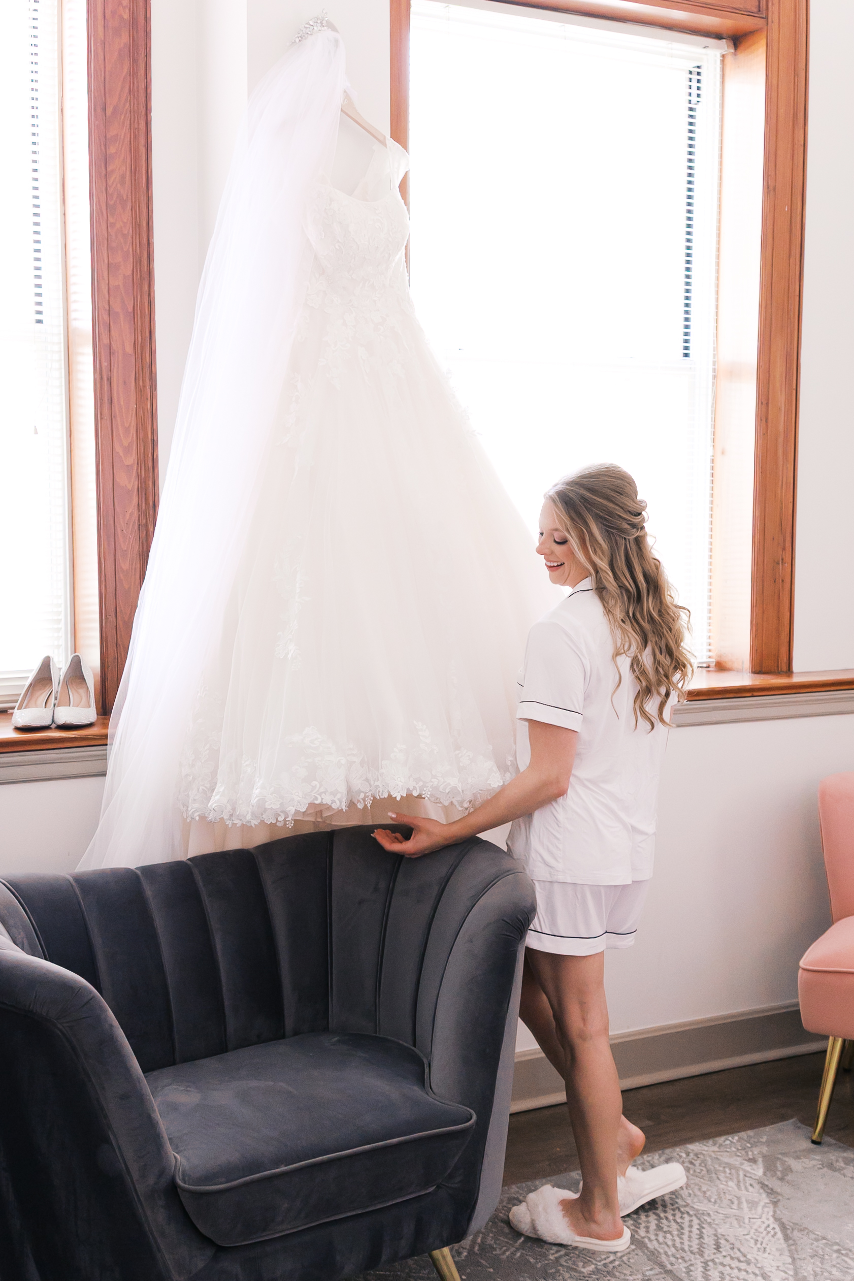 Bride in white getting-ready pajamas admiring her lace wedding dress hanging in a bright bridal suite