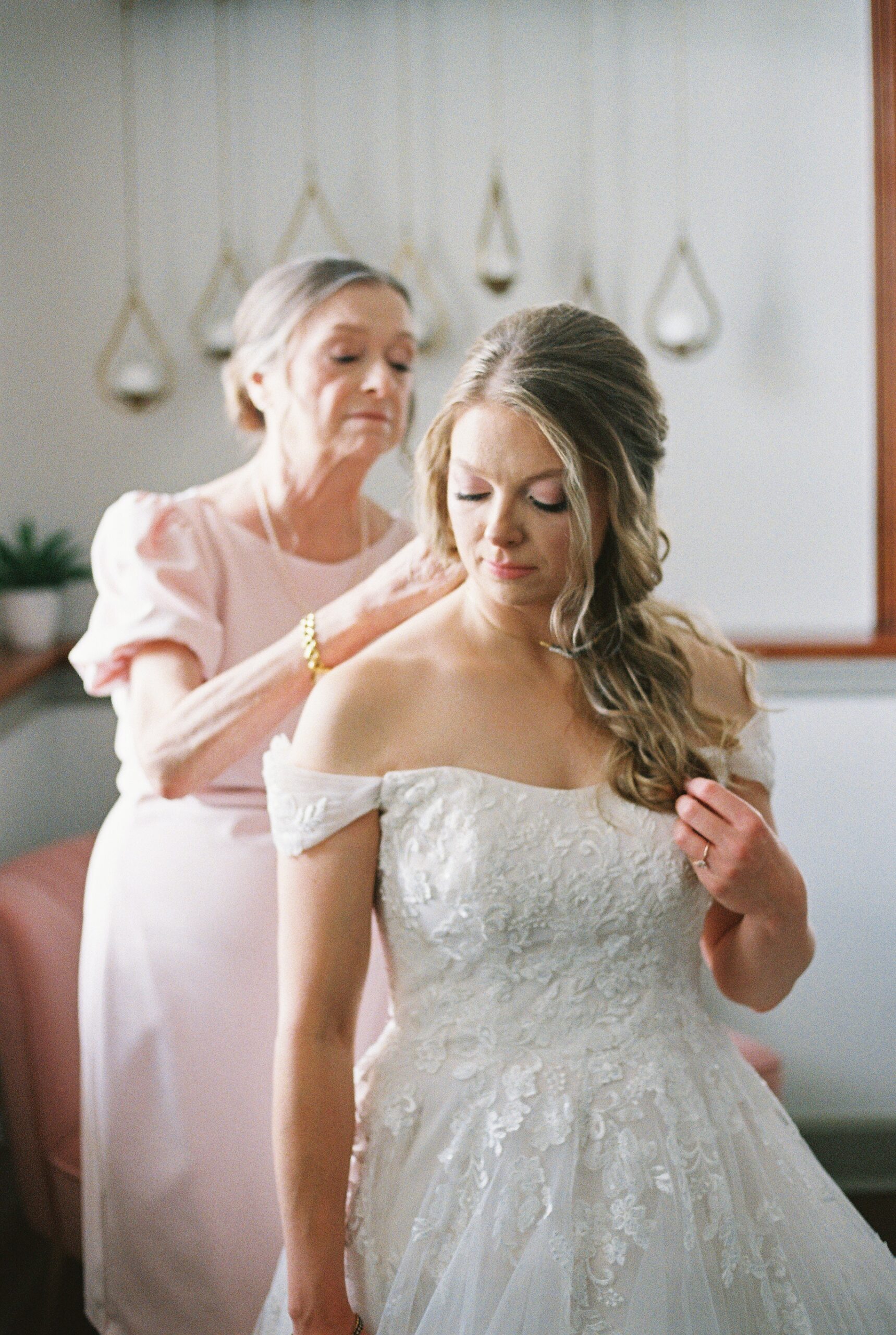 Mother fastening the bride's off-the-shoulder lace wedding gown during wedding preparations.