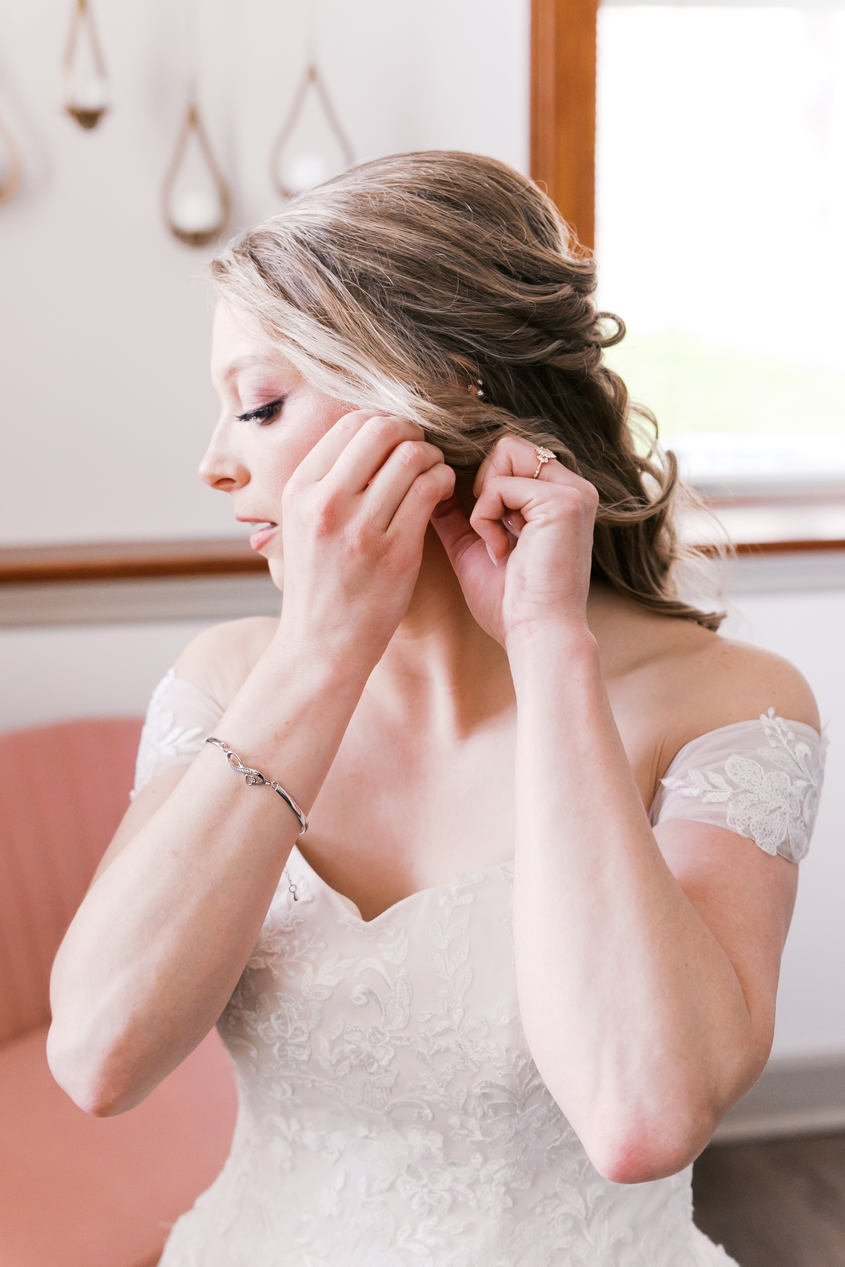 Bride fastening an earring while getting ready in an off-the-shoulder lace wedding dress.