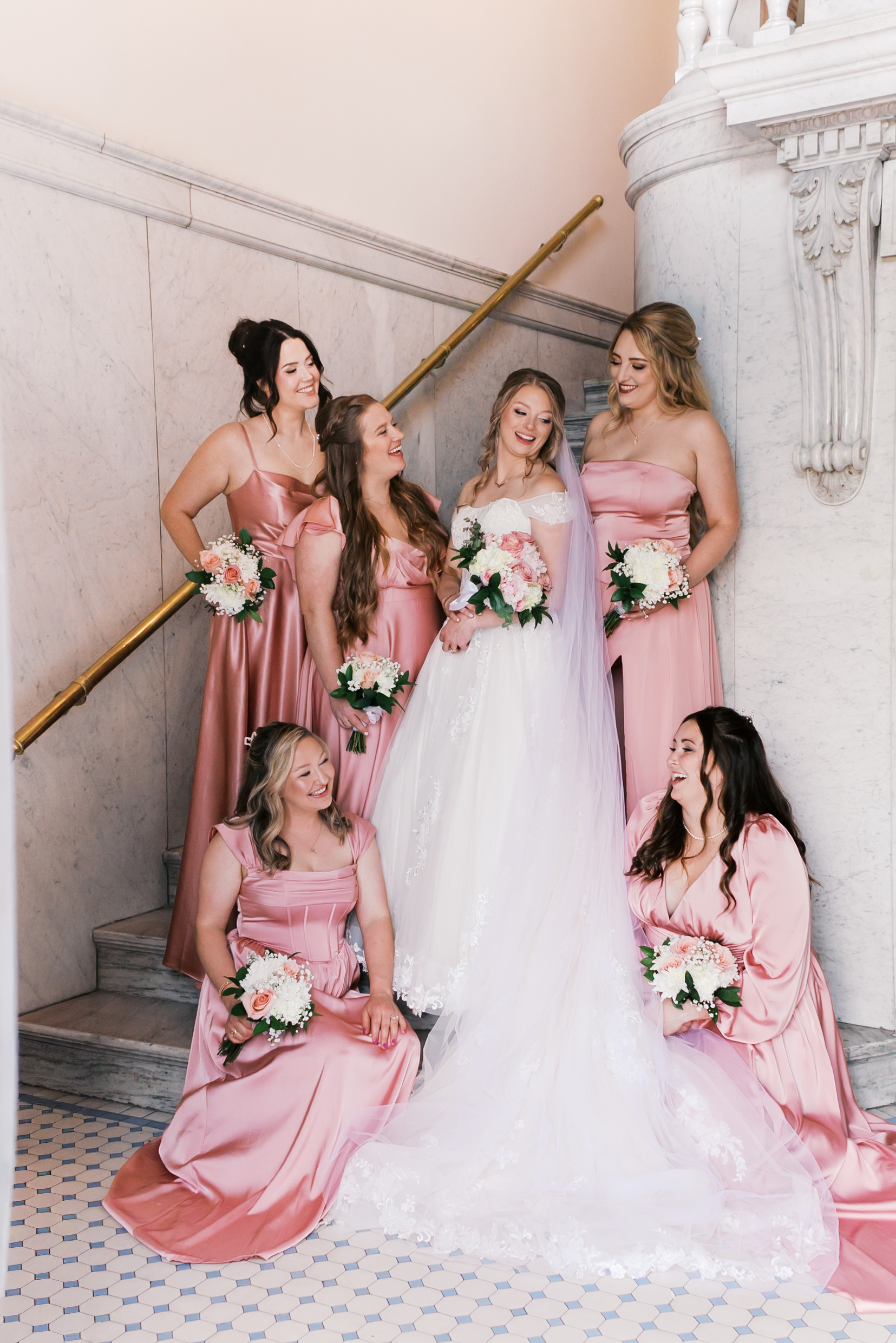 Bride with bridesmaids in blush pink dresses posing on a marble staircase with bouquets.