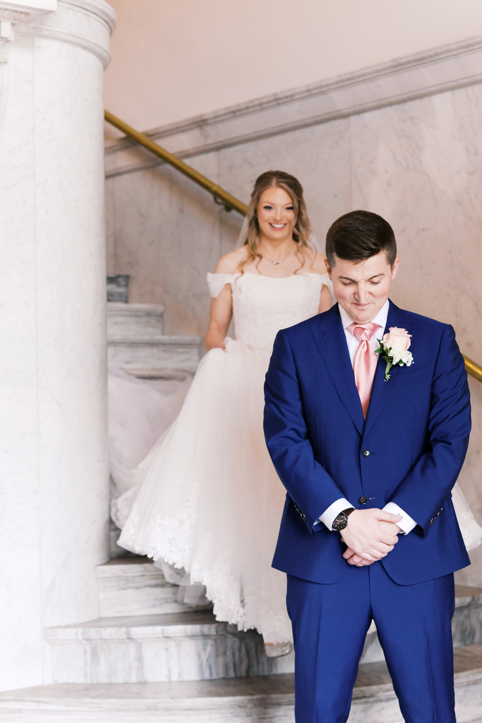 Groom waiting for the first look while the bride approaches from behind on a marble staircase.