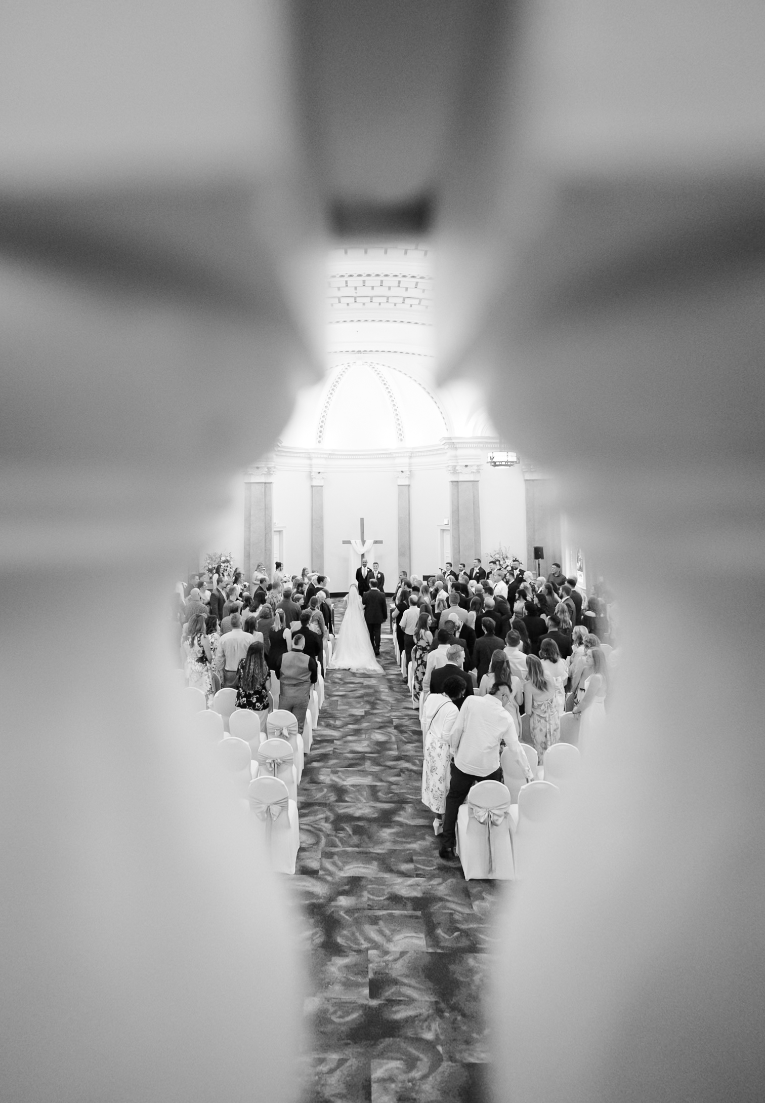 Black and white view of a wedding ceremony through a narrow architectural opening.