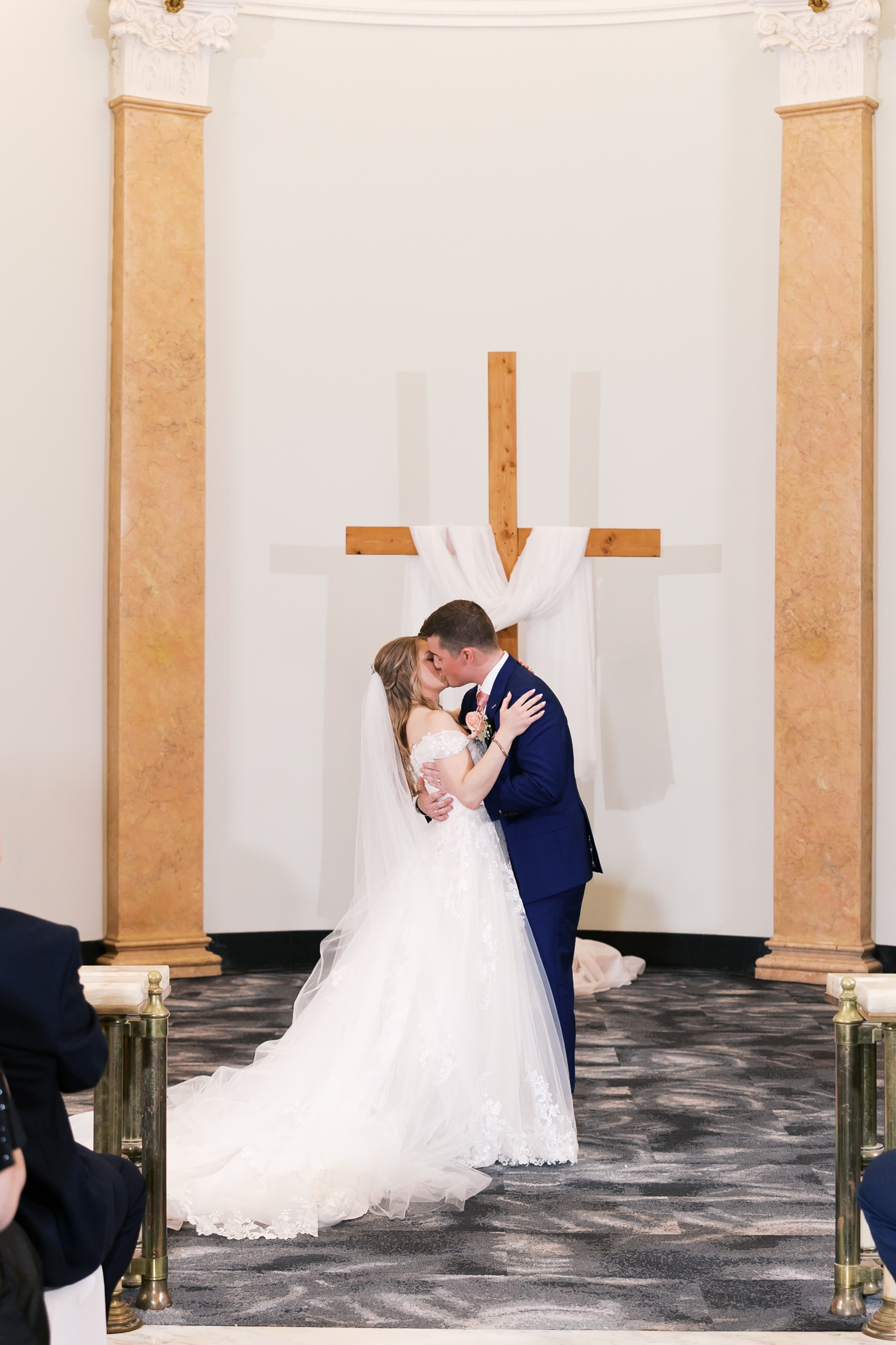 Bride and groom sharing their first kiss at the altar beneath a wooden cross.