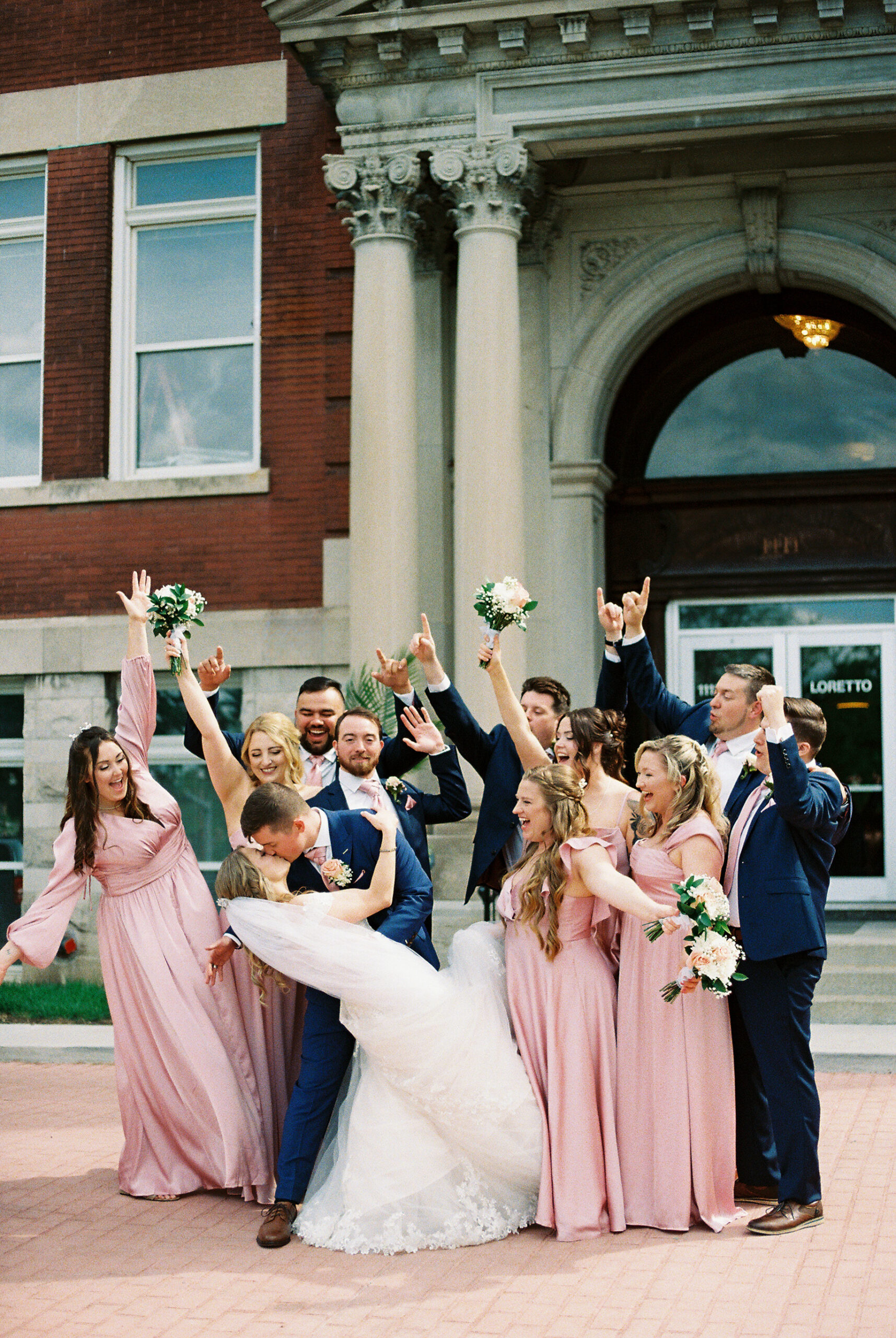 Bride and groom kiss while bridal party in blush dresses and navy suits celebrates outside historic wedding venue.
