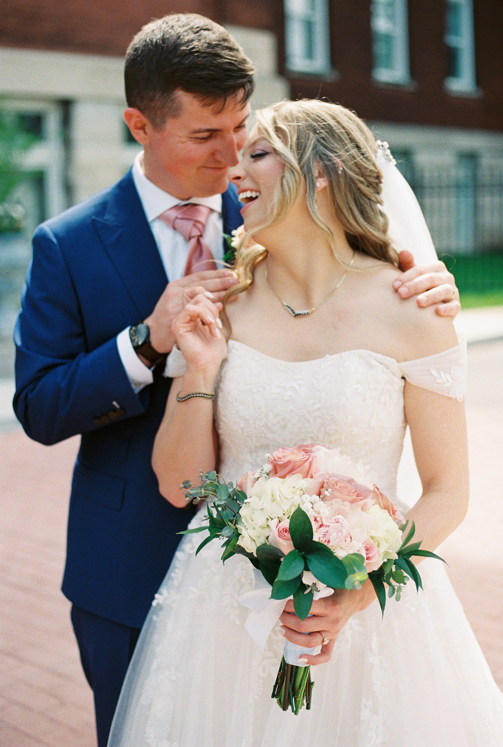Bride and groom embrace outdoors, holding a blush and white rose bouquet during wedding portraits.