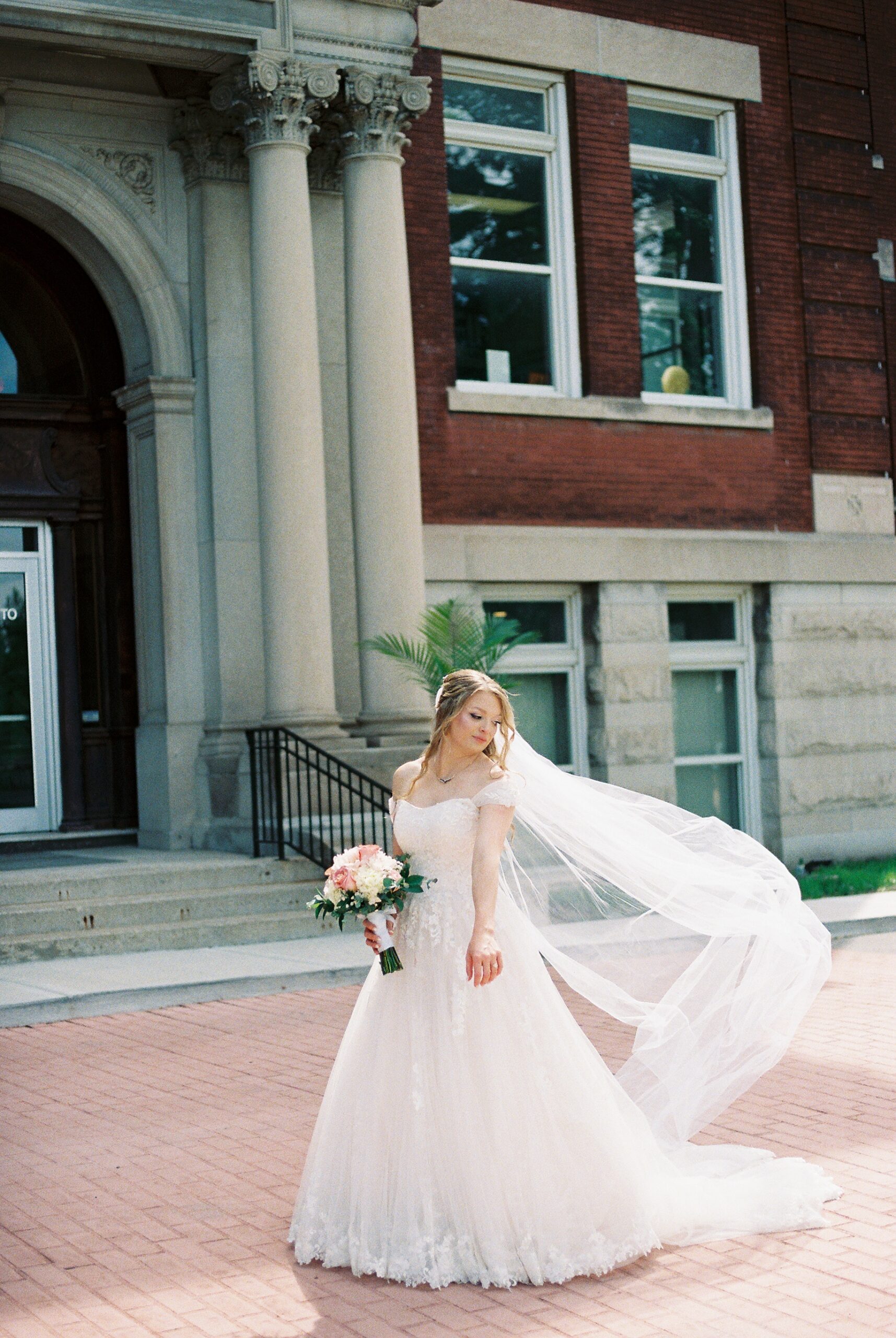 Bride poses outside historic wedding venue as her cathedral veil flows in the breeze, holding a blush and white bouquet.