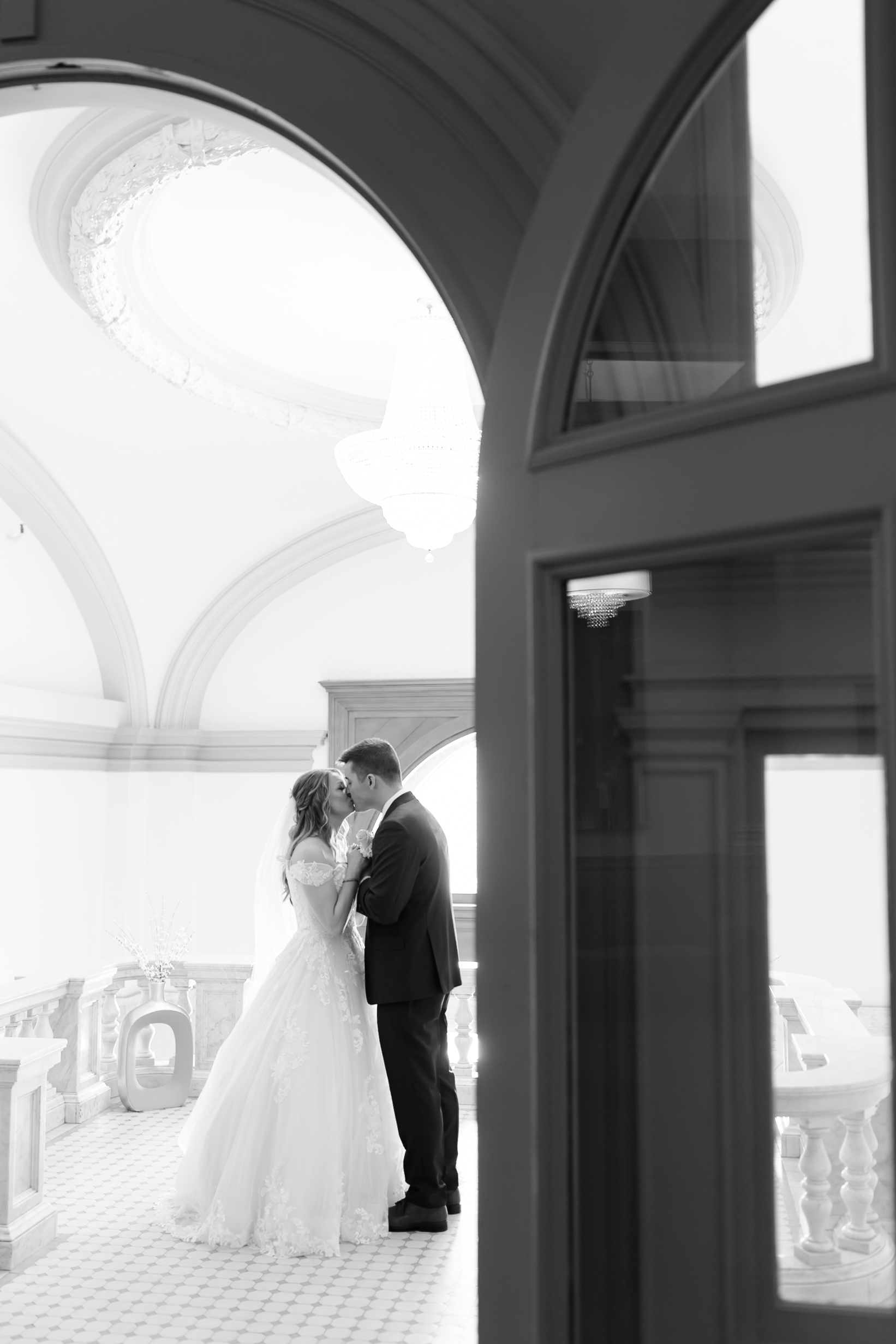 Bride and groom share a kiss beneath arched architecture in a bright marble hallway during wedding portraits.