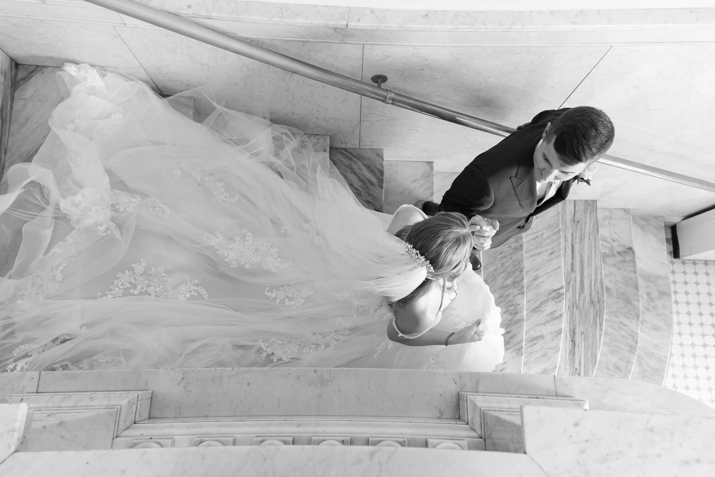 Overhead view of bride and groom holding hands on a marble staircase, showcasing the bride's flowing lace train.